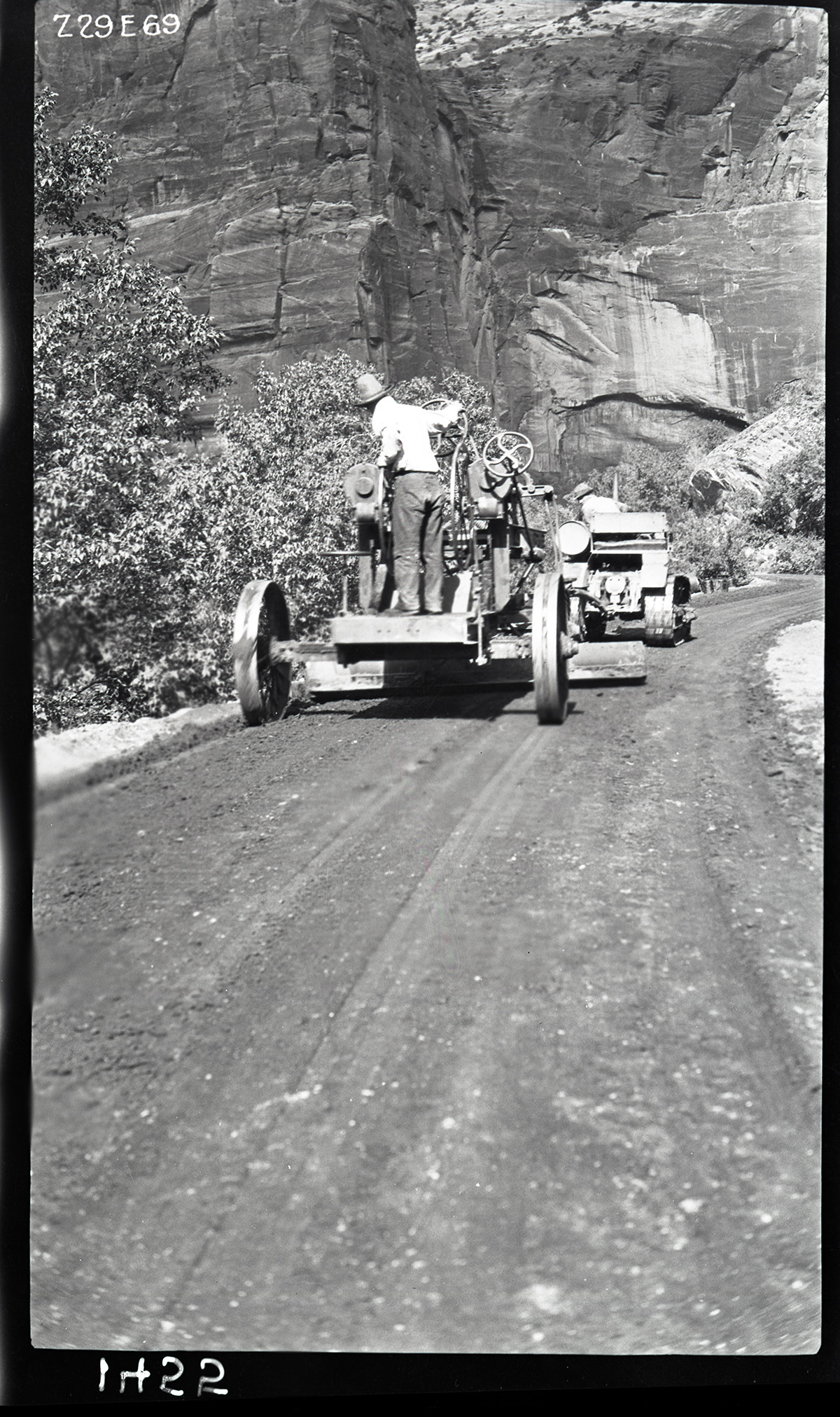 Worker driving a road grader following another worker on a caterpillar tractor on the floor of the valley road spreading paving materials. Labeled 'experimental road paving.'