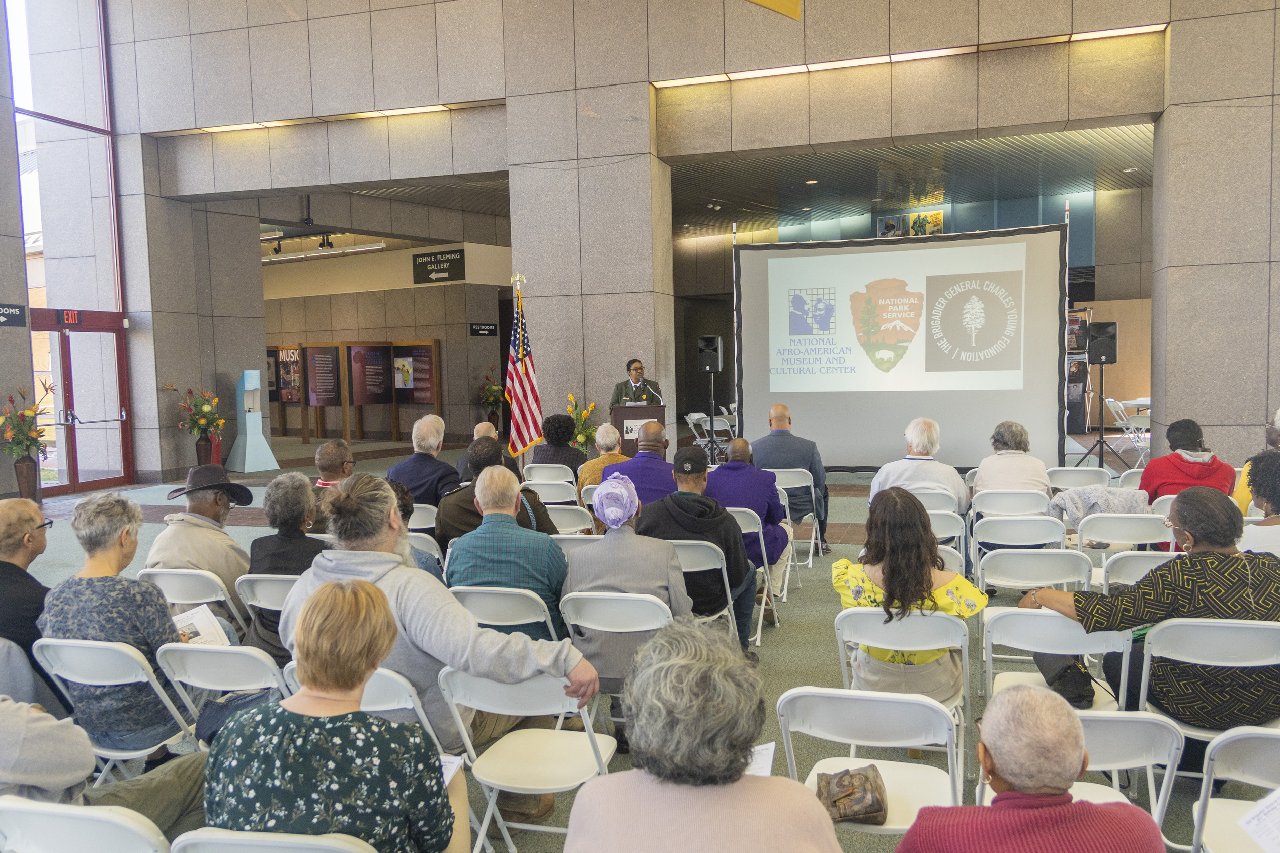 A lady in a green ranger uniform standing behind a wooden podium speaking into a microphone atop the podium. 