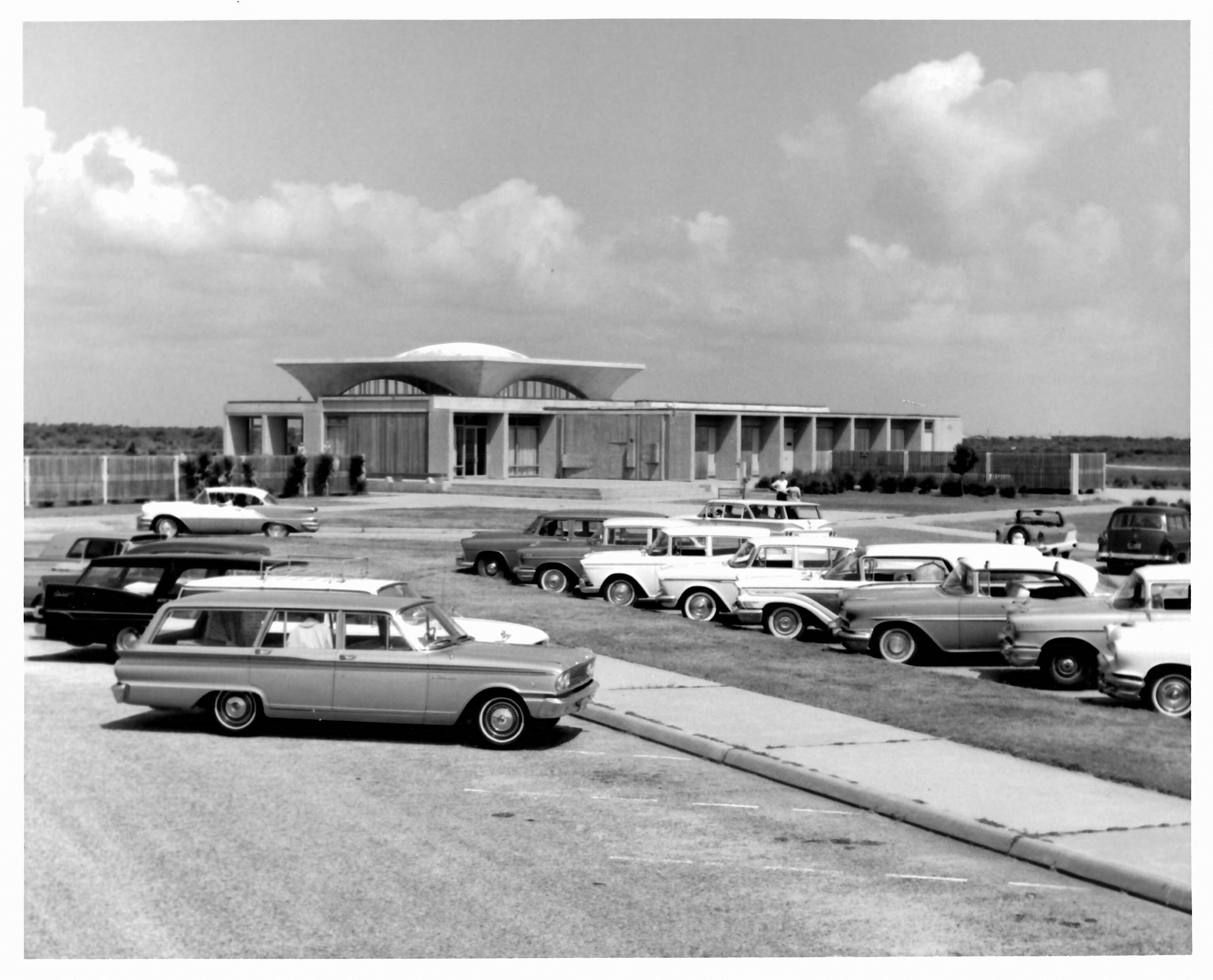 Black and white photo of 1960s-era cars parked in parking lot in front of visitor center. Visitor center seen in background. Billowy clouds seen in sky.