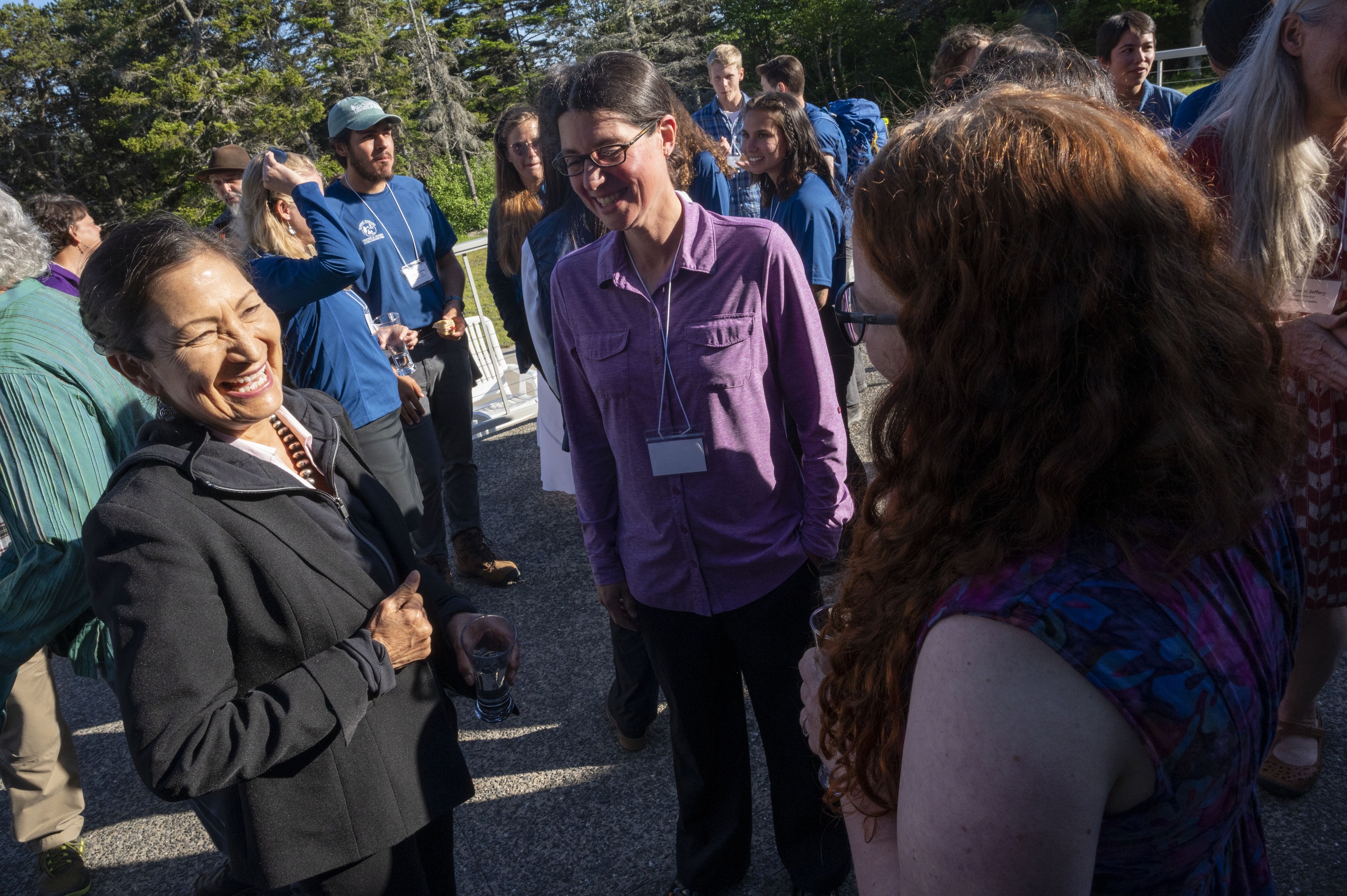 Woman in a dark jacket laughs and smiles wide with two women surrounded by clusters of other people engaged in separate conversation.