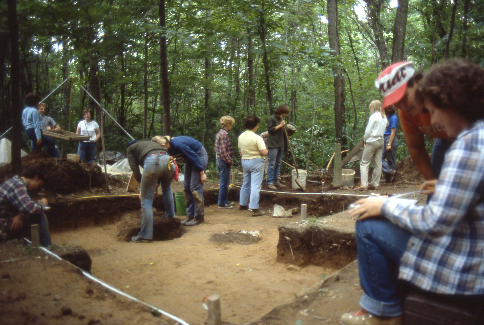 A dozen people sit and stand in and around a brown, angular archeological excavation in a forest.