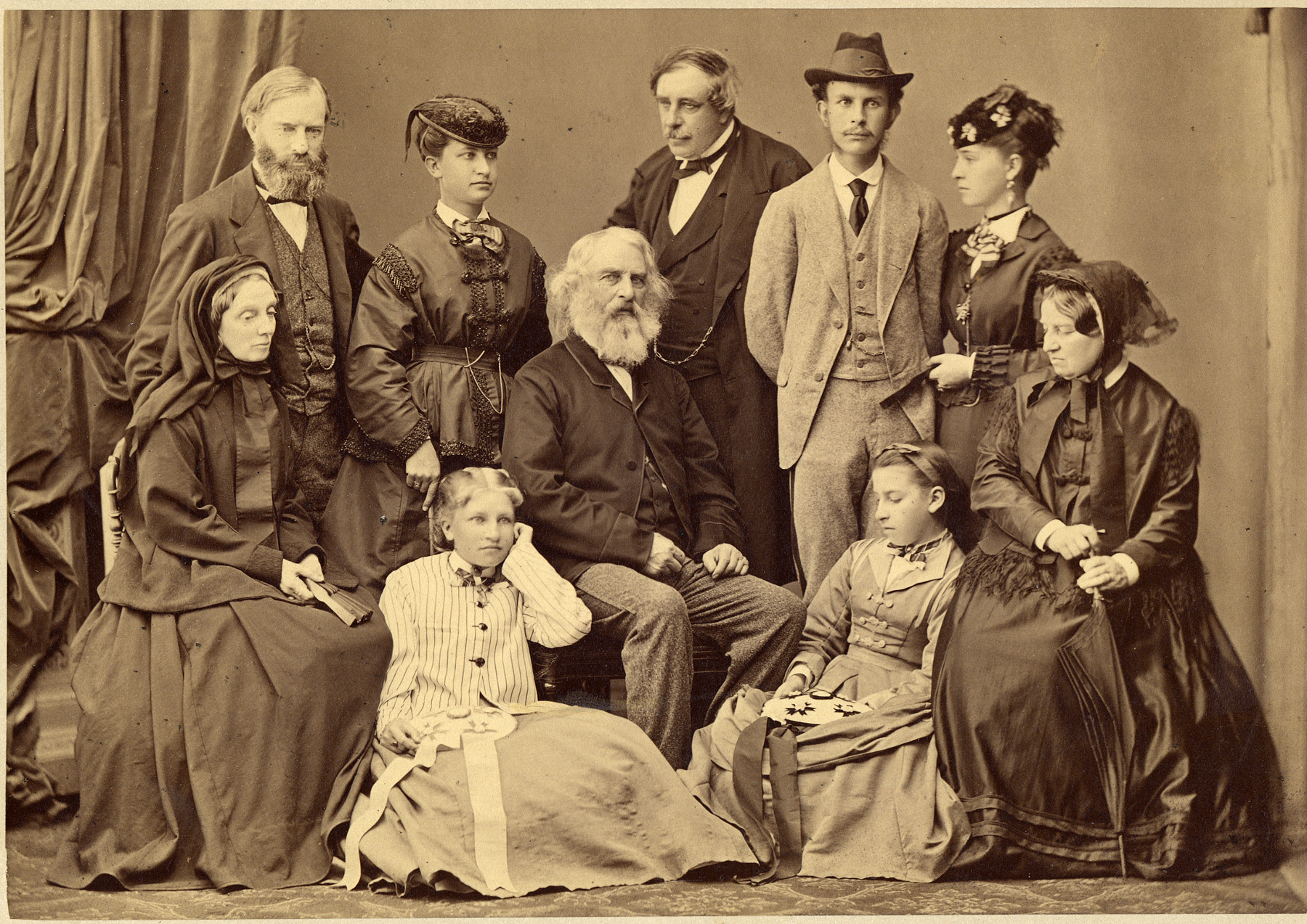 Sepia tone photograph of Longfellow family in photographer's studio. Three men and two women stand in back row. Henry Longfellow seated at chair in center. Two older women sit in chairs at left and right, two teenage girls sit on floor.
