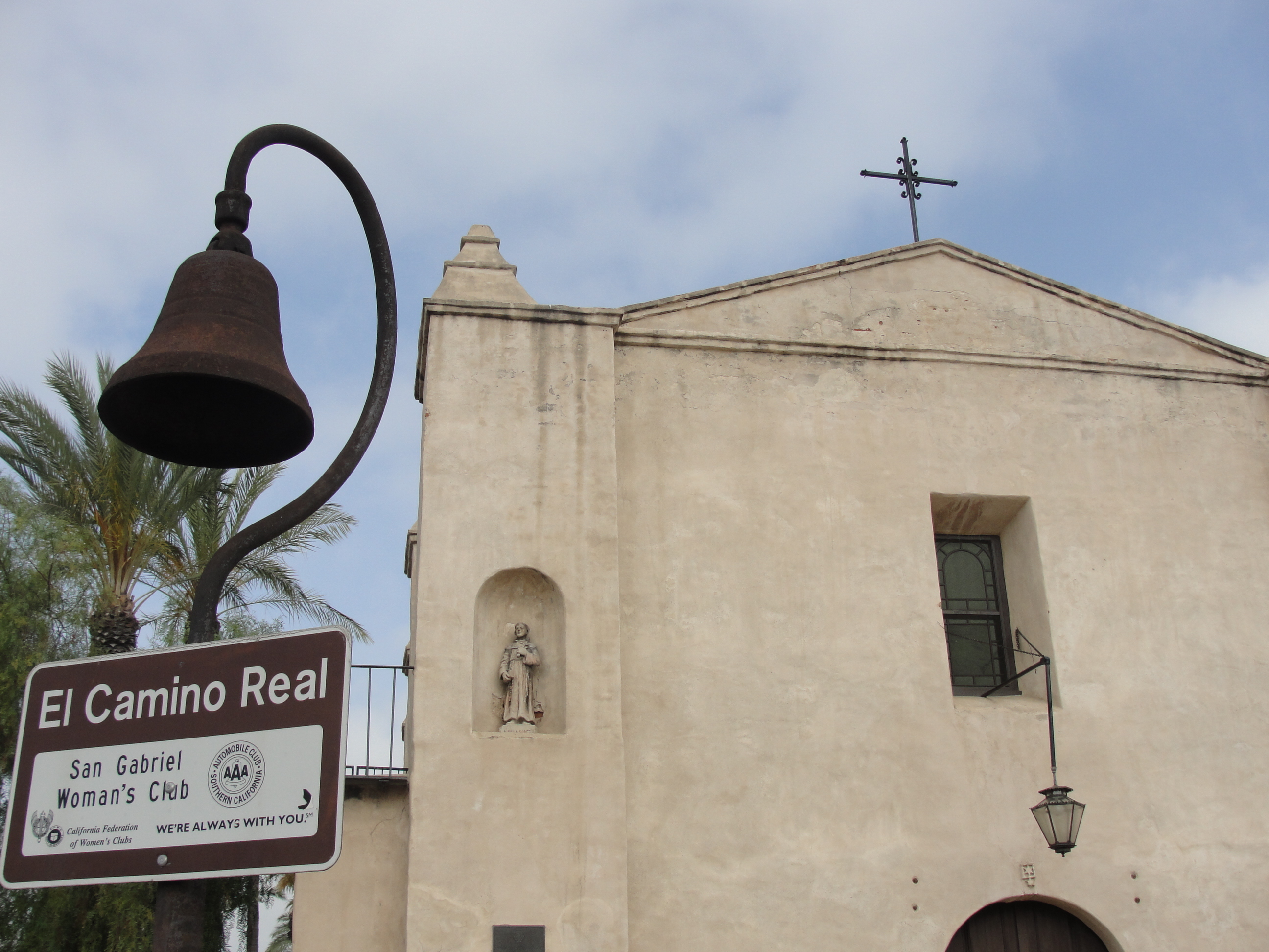 The front of a white mission building with a cross on the roof and a bell in front for El Camino Real