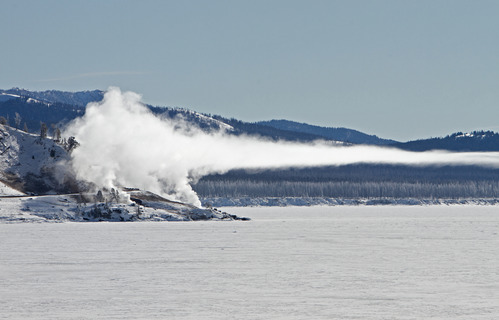 Steam rises from thermal features next to a frozen lake and the steam drifts out over the lake in a flat layer.