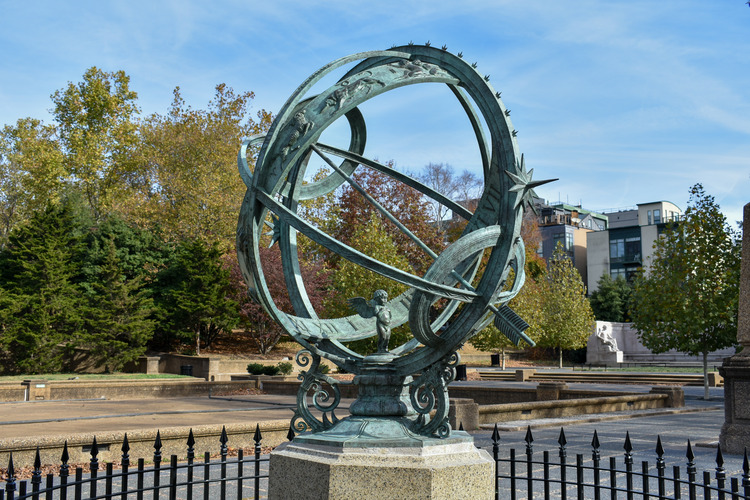 A green-tinted armillary sphere sculpture surrounded by bushes and pointed iron fence. It stands in front of fall trees and buildings in the distance.