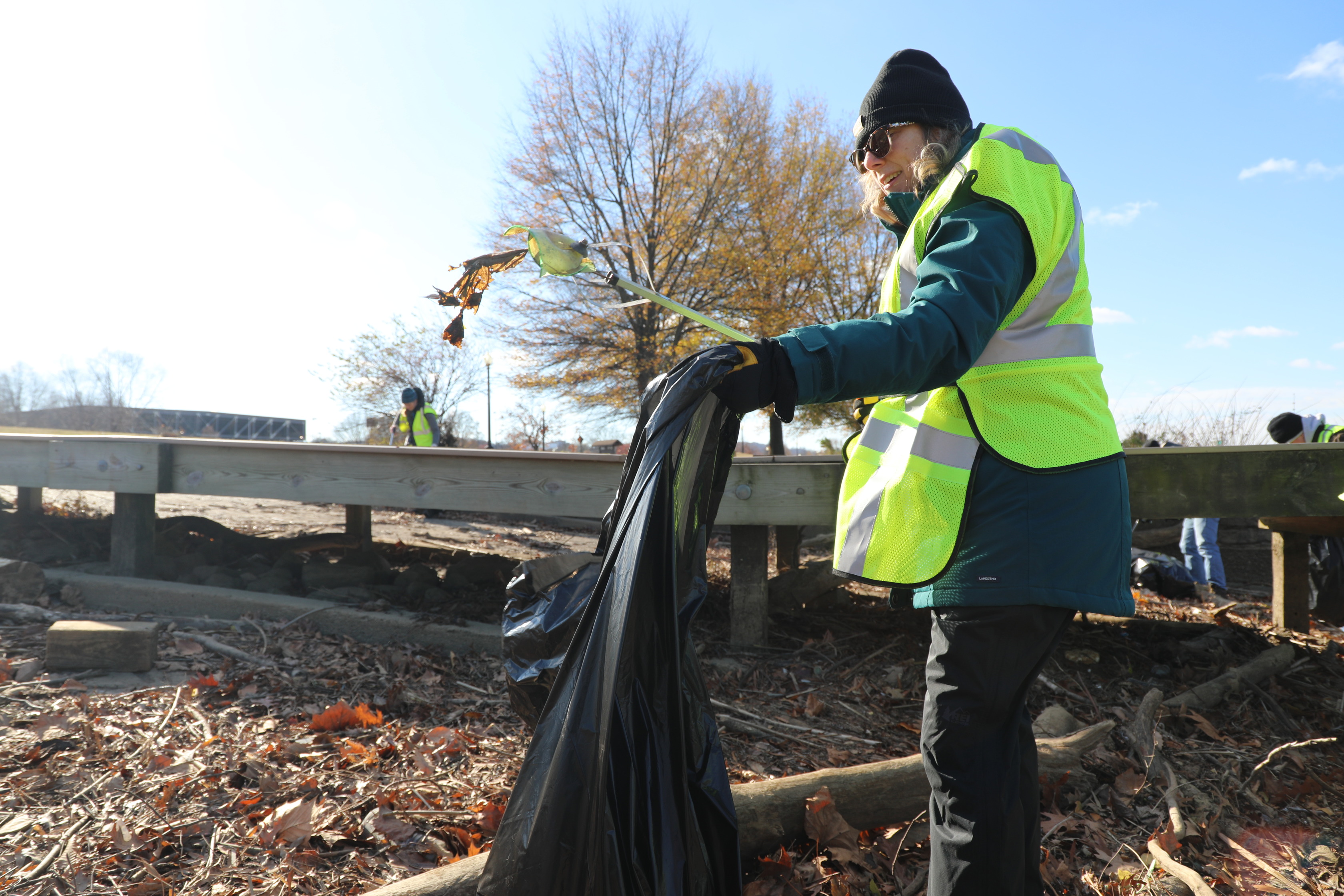 A woman, dressed in a dark winter coat, black beanie, sunglasses, and a bright yellow reflective safety vest, is actively picking up trash. She is holding a litter picker tool in her right hand, using it to place debris into a large black trash bag held in her left hand. The ground is covered with fallen leaves and branches. In the background, there is a wooden structure or boardwalk, and a few other individuals in similar safety vests are visible. The sky is bright and clear, and a deciduous tree with bare branches stands in the middle distance.