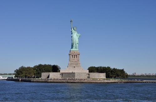 Patina-colored copper colossal statue of a woman with a crown and uplifted right arm stands on a granite pedestal on an island. 