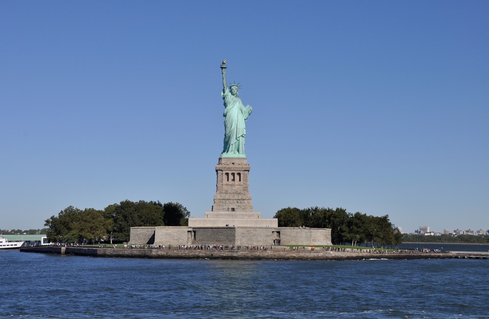 Patina-colored copper colossal statue of a woman with a crown and uplifted right arm stands on a granite pedestal on an island. 