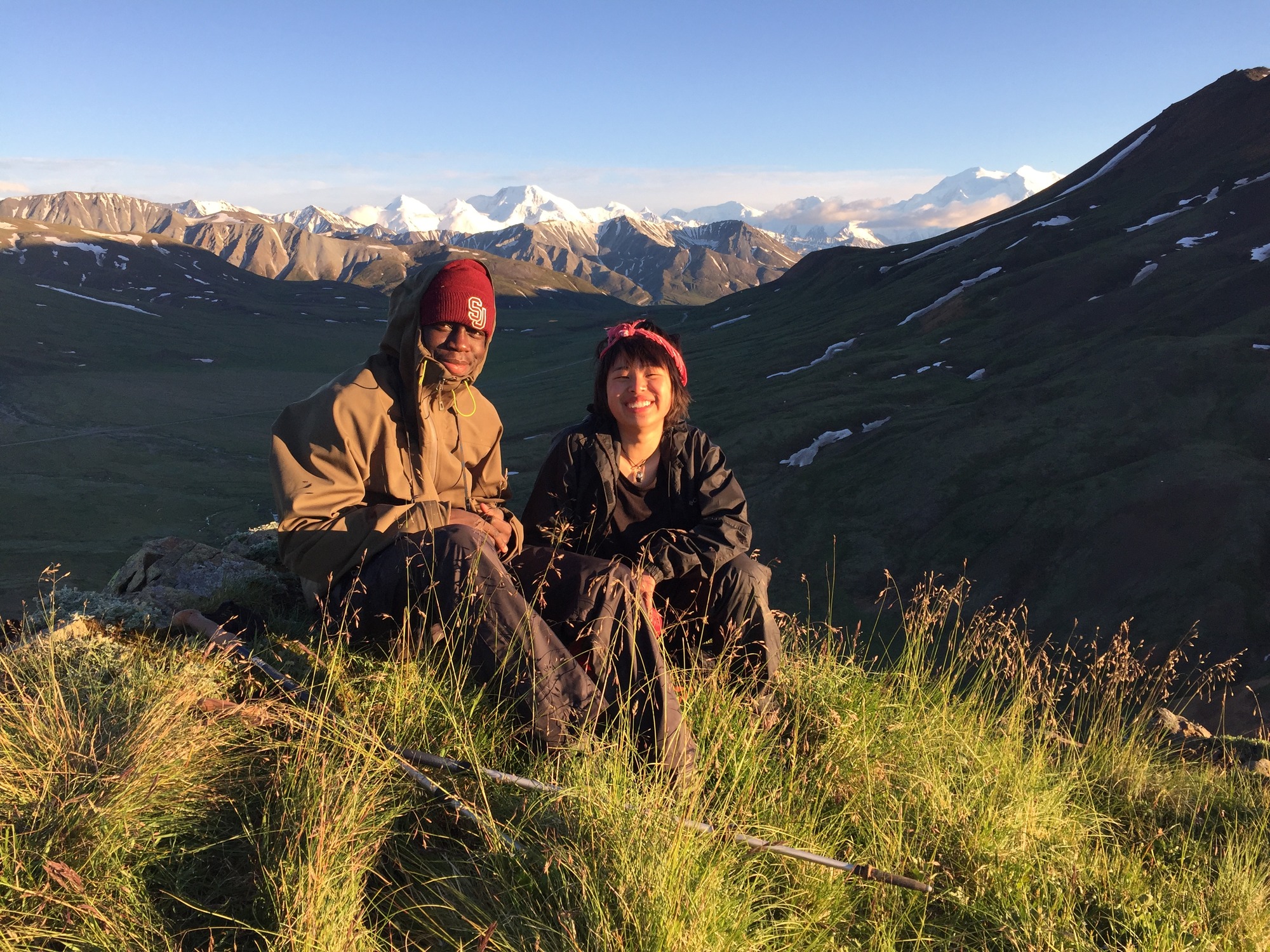 two people sitting on a mountaintop looking out across a valley and distant, snowy mountains