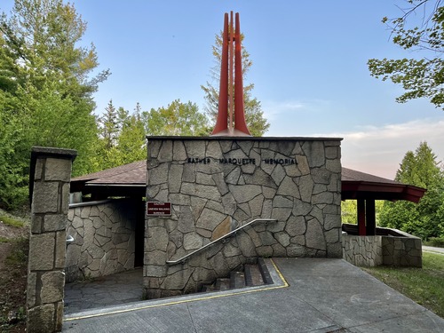 Lettering reading "Father Marquette Memorial on a stone wall above a staircase leading into a stone memorial structure with a pointed red roof
