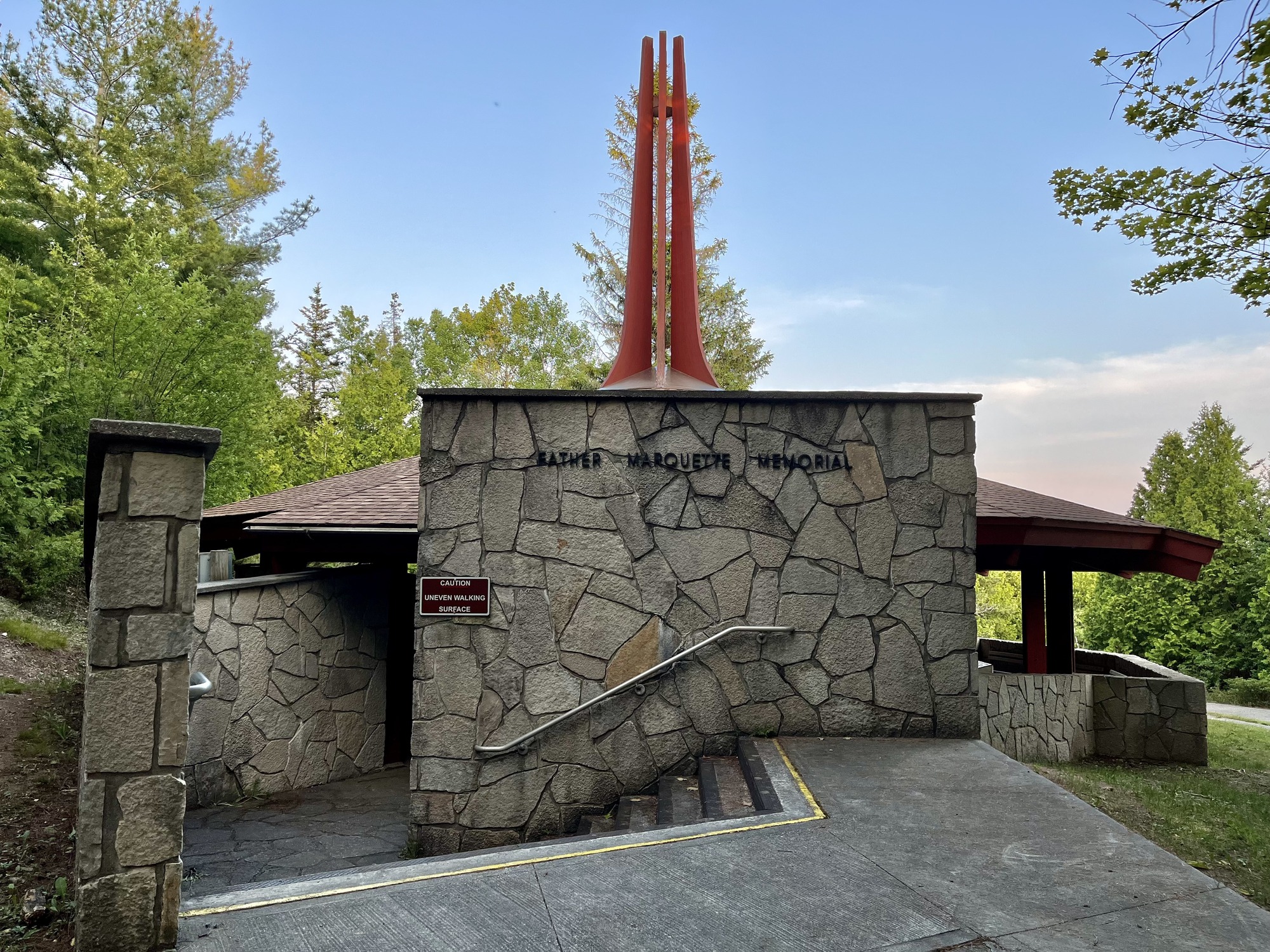 Lettering reading "Father Marquette Memorial on a stone wall above a staircase leading into a stone memorial structure with a pointed red roof