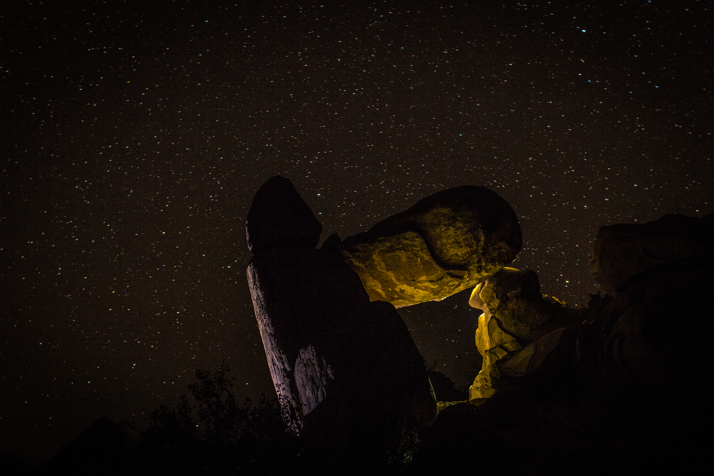 A balanced rock formation is dimly lit under a dark, starry night sky.
