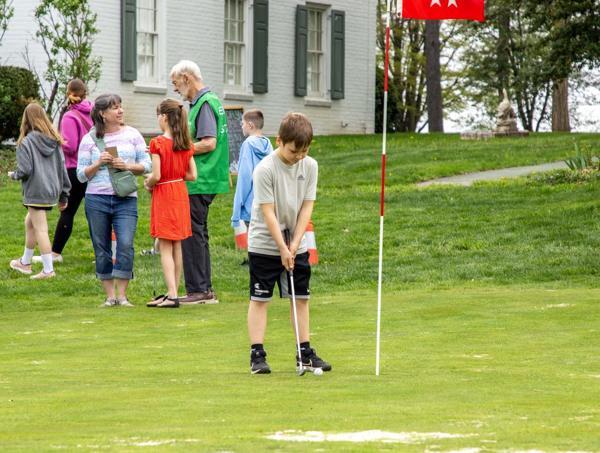 A young visitor with a putter stands on a green putting green next to a red flag, with volunteers, visitors, and the white brick Eisenhower home in the background. 