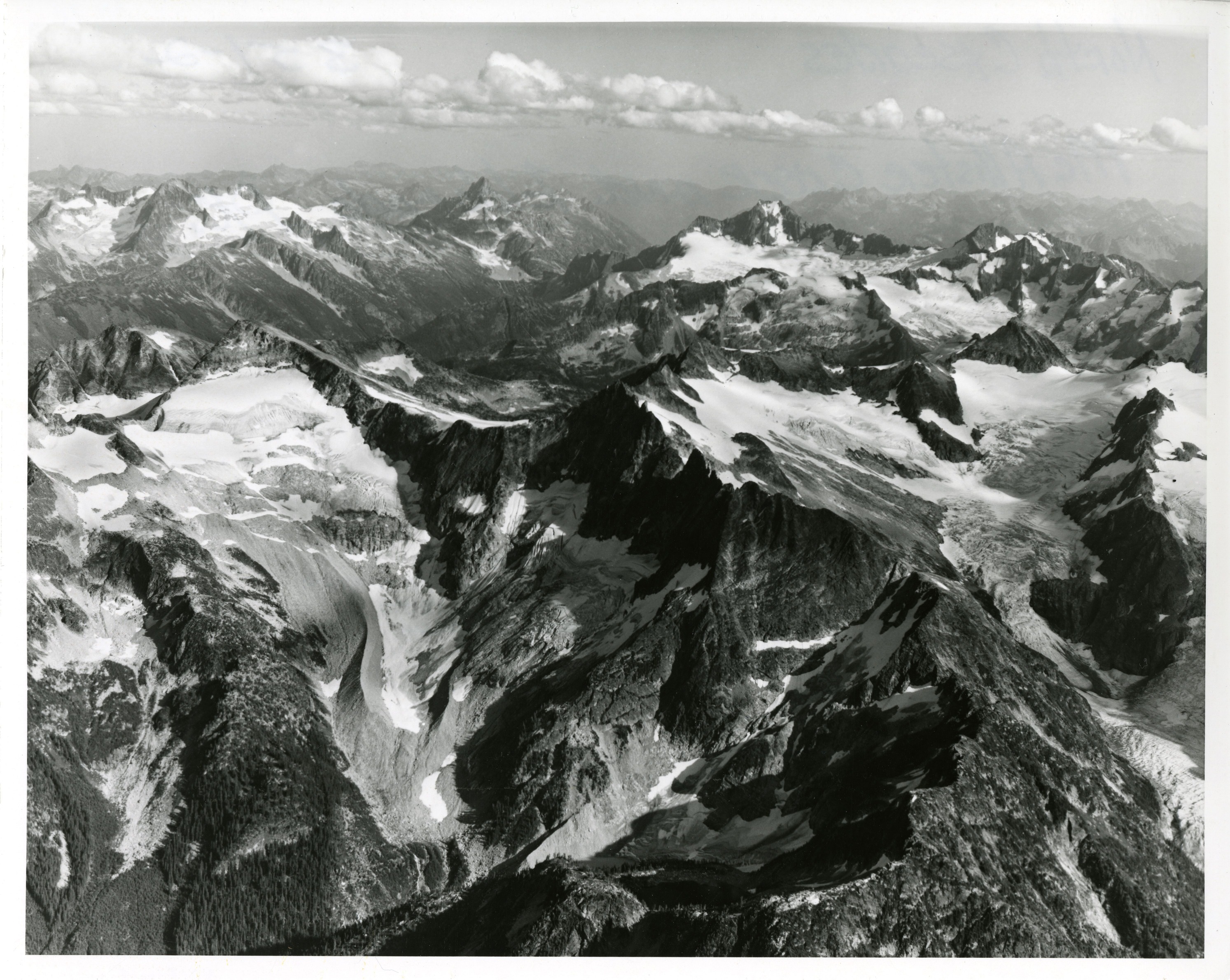 Aerial view of a mountain ridge. The peaks of the mountains are covered in glaciers. More mountain peaks can be seen in the background.