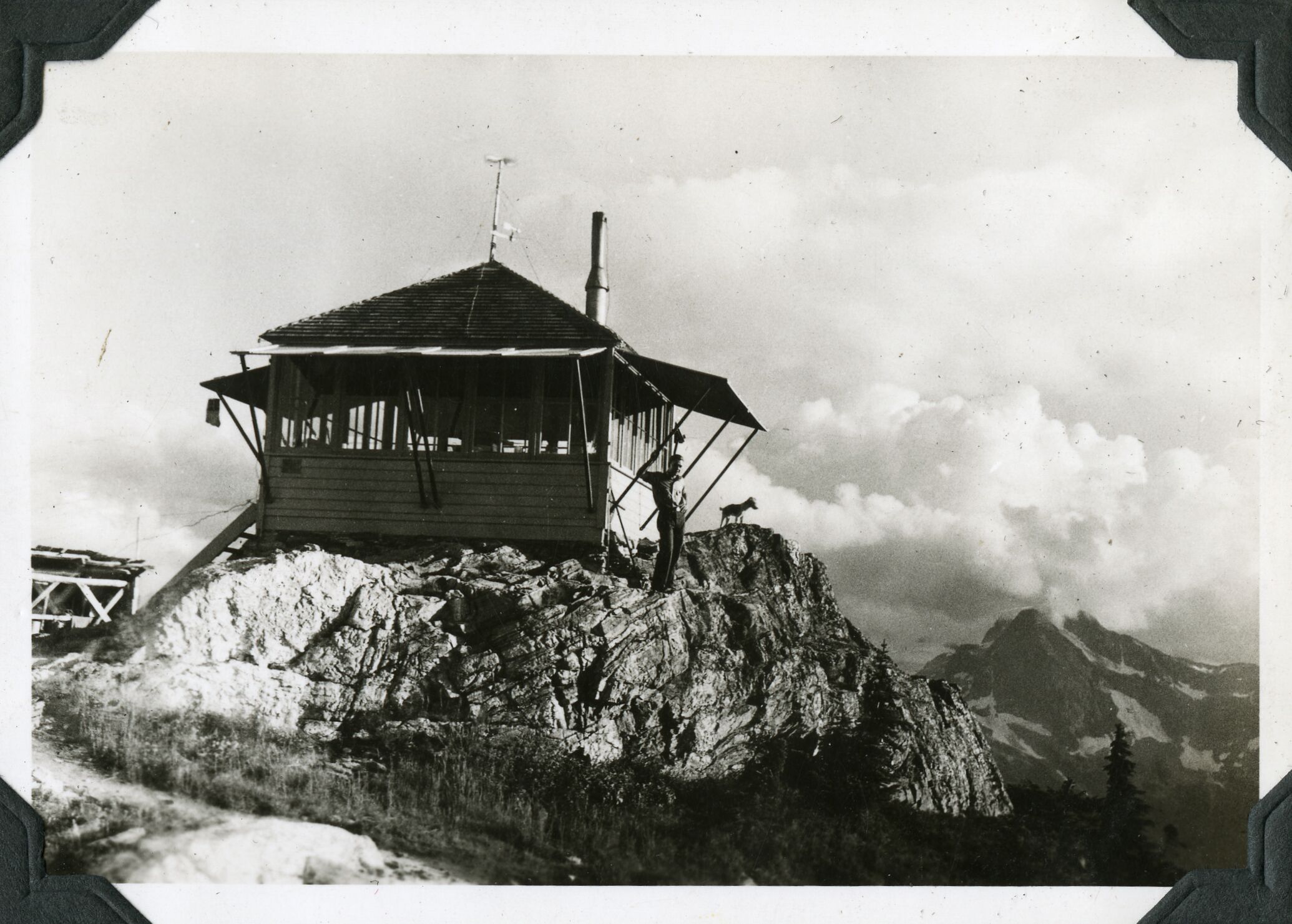 A man and a dog stand against a wooden structure on a rocky outcropping.