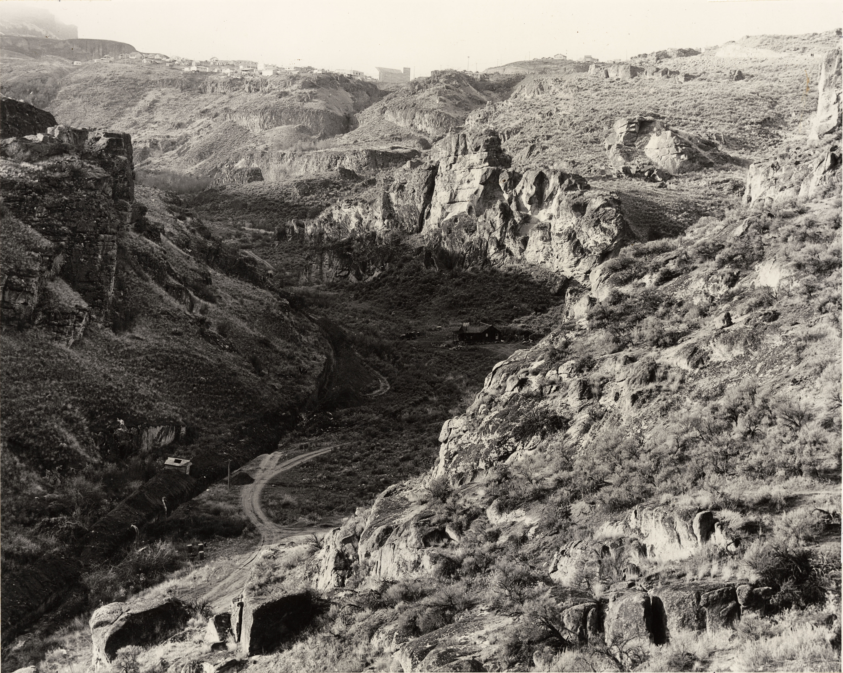Black and white photograph of a canyon with large rock outcrops. A road runs through the canyon and buildings can be seen on the hill above.