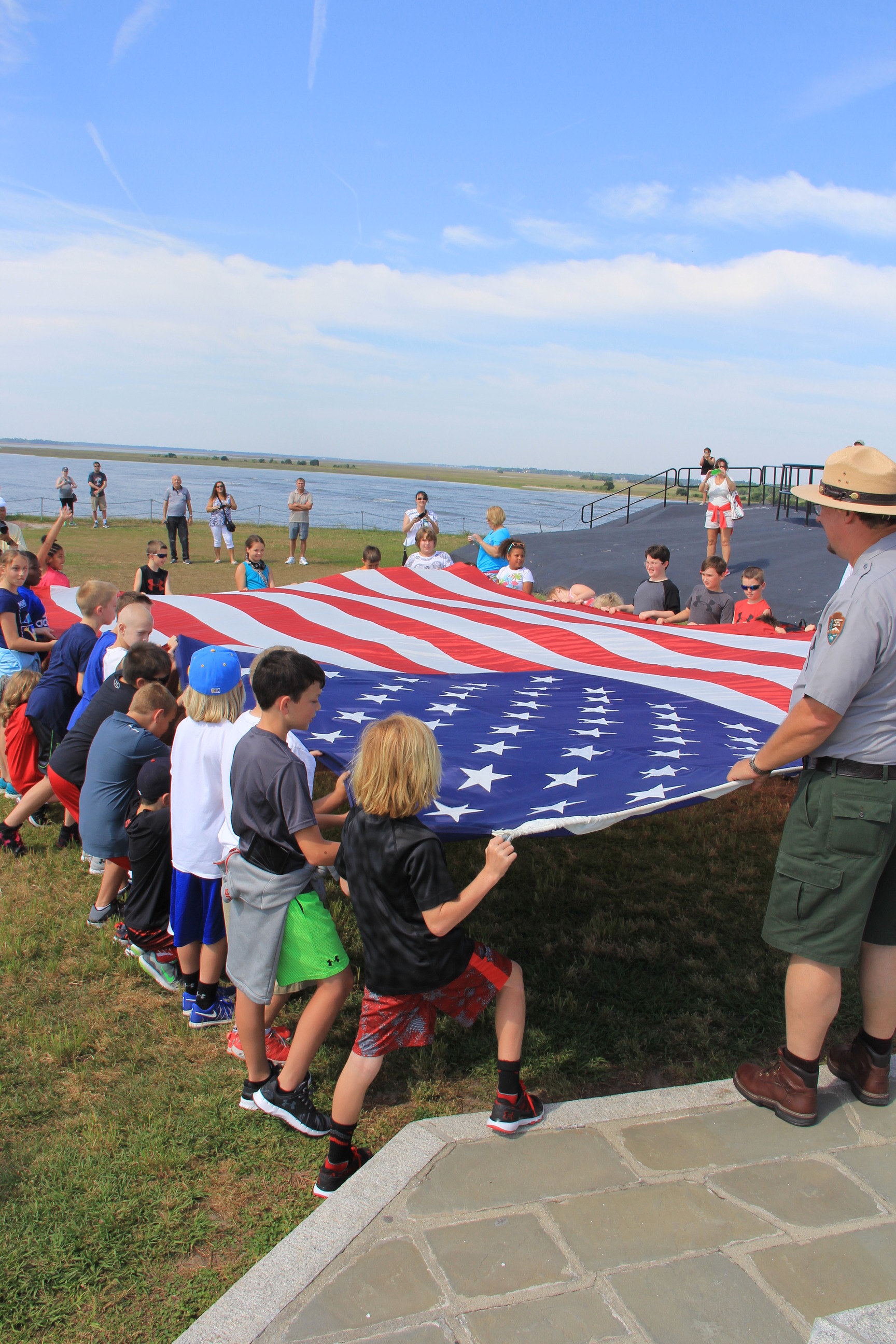 A park ranger and a couple of dozen kids hold a large American flag on grass near a large body of water.
