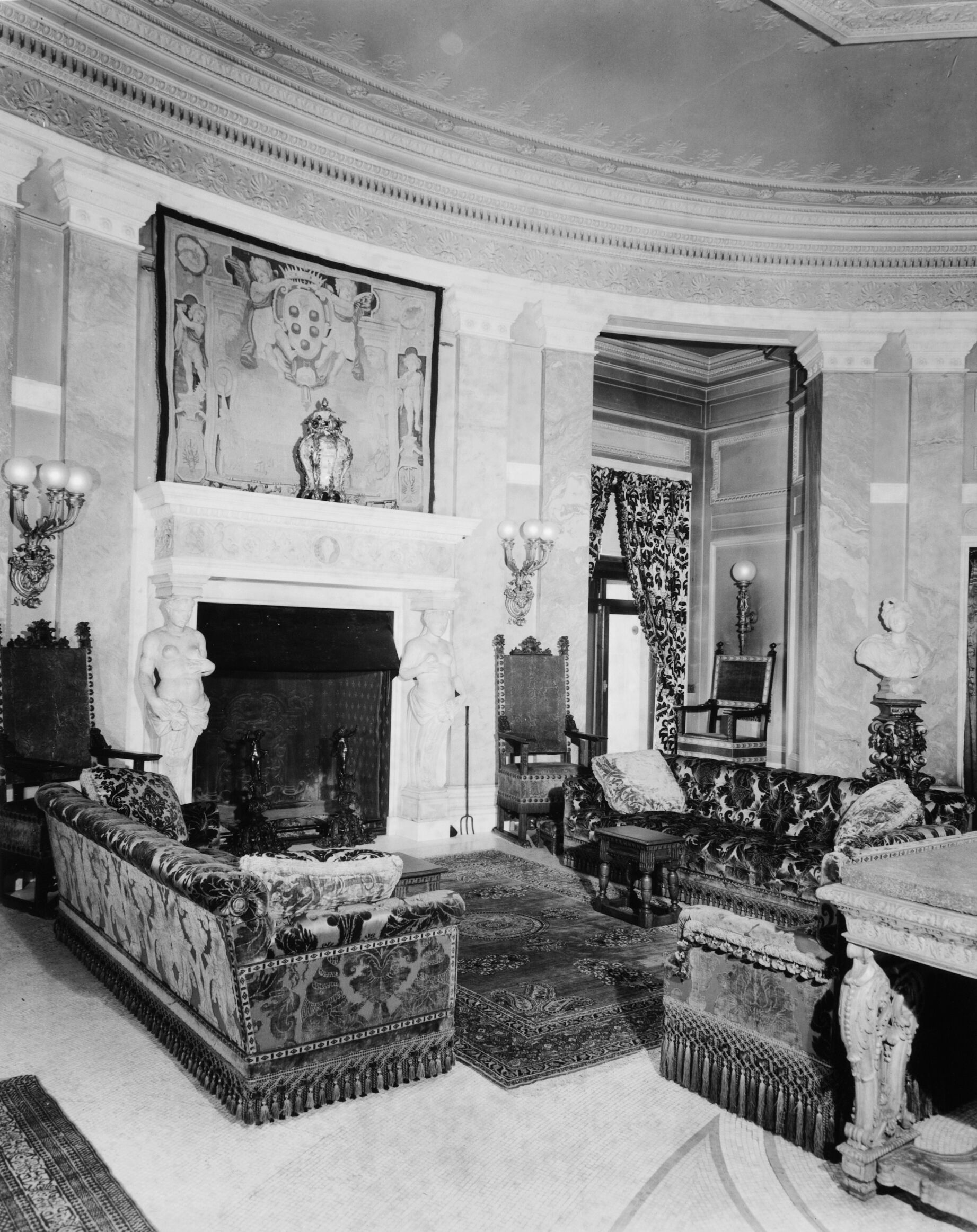 An oval shaped hall with a tapestry above a large fireplace and sofas.