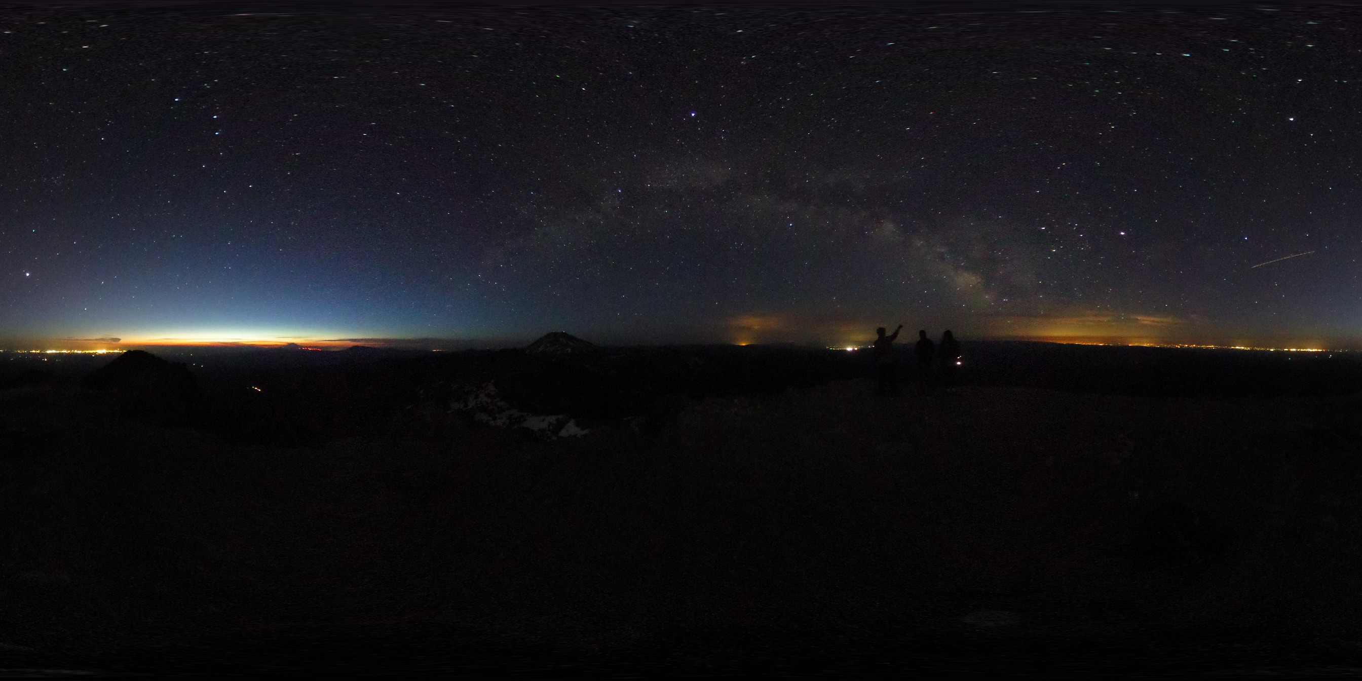 A 360 color photo of a rocky summit of a mountain at night. Three hikers stand beneath a starry sky with a cloud-like arch across the sky (Milky Way). 