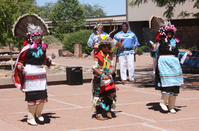 Colorfully dressed Zuni dancers with singers and drummers in the back, in a plaza with buildings and trees in the background.