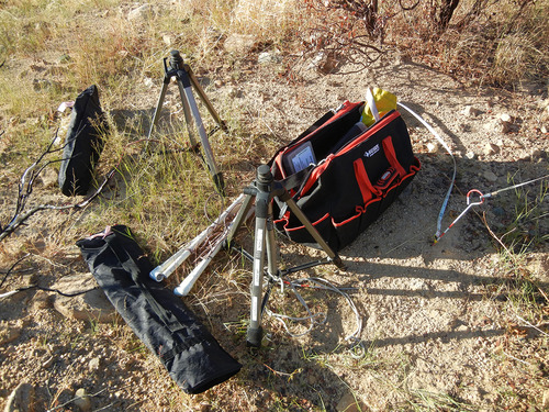 Photo of bags, tripods, measuring tape, and pin sitting on bare ground