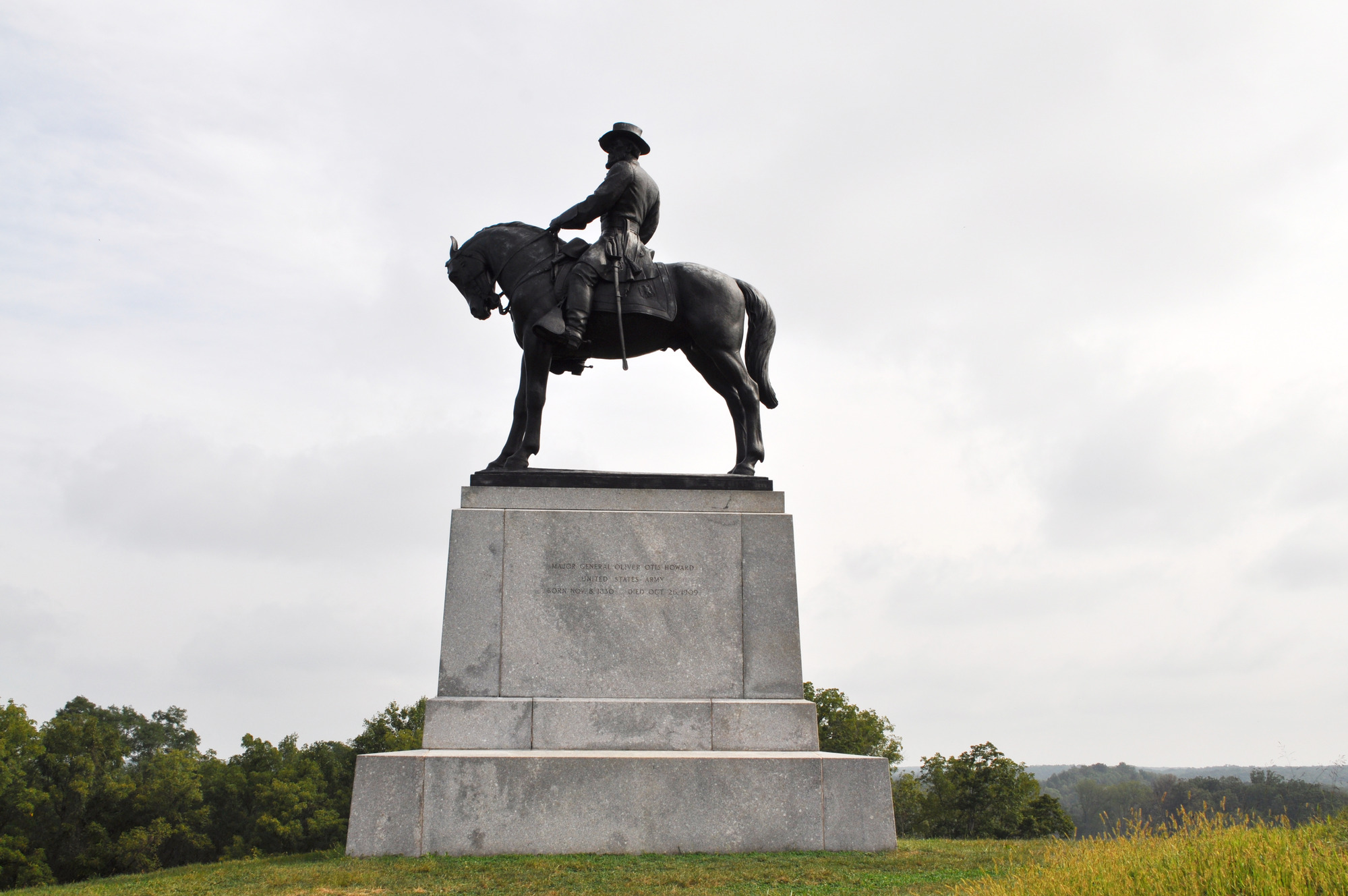 A statue of a man on a horse with its head bowed, facing left, surrounded by low grass, hazy trees, and overcast sky. 