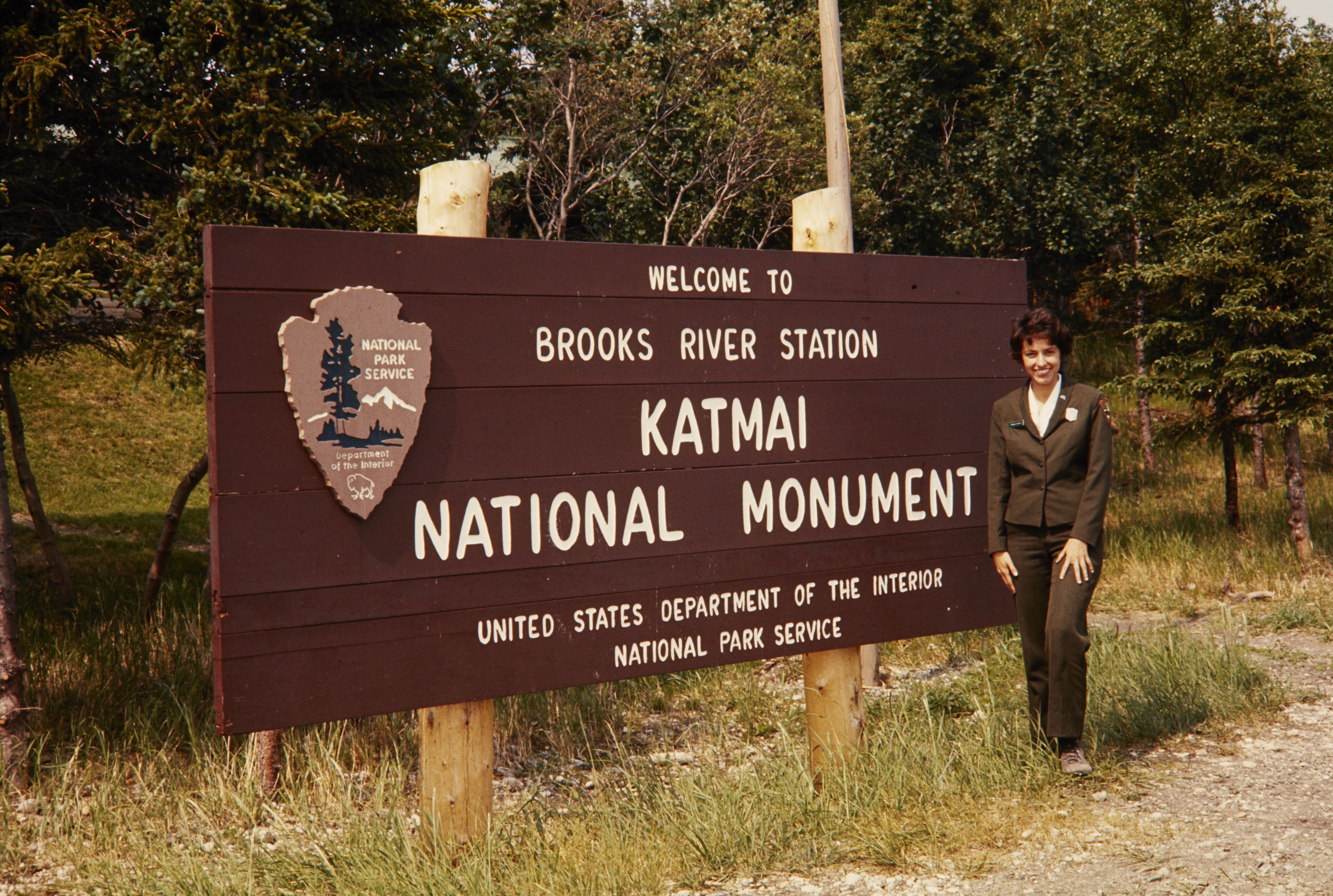 Bonnie Koploy in her NPS uniform stands by the sign for Katmai National Monument.