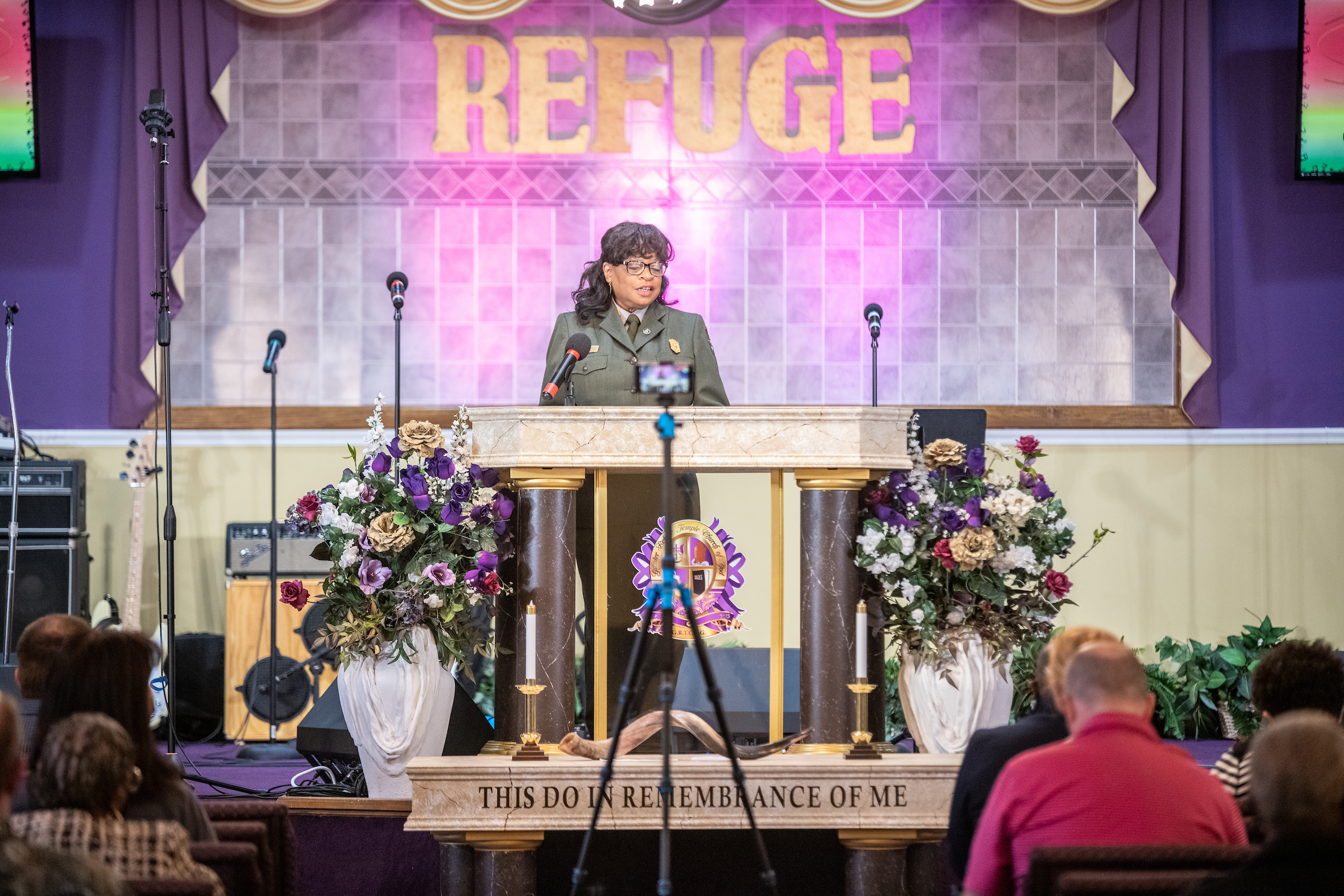 Superintendent Deanna Mitchell can be seen in the middle of the picture giving a speech to a crowd in the foreground. Purple drapes, a beige wall, and golden letters spelling out the word, "REFUGE" can be seen in the background.