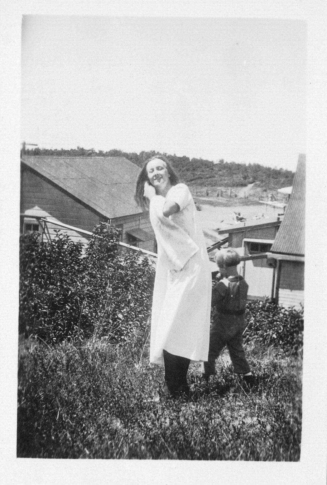 A black and white image of a woman and a child outside on a lawn above a community. The woman is located in the center of the image. She is wearing a short-sleeve dress. She is smiling at the camera. Her left hand is reaching across her chest adjusting her hair on the right side of her head. She has a scarf or jacket draped on her left arm. Her right arm is holding the hand of the boy behind her. The backside of the boy is visible. He is wearing a long sleeve shirt and pants and is facing towards the left side of the image. The woman and boy are standing in a grassy area uphill from two houses in the background.