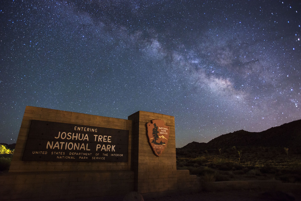 Night sky scene shows the Milky Way galaxy arching over the "entering Joshua Tree National Park" entrance sign.