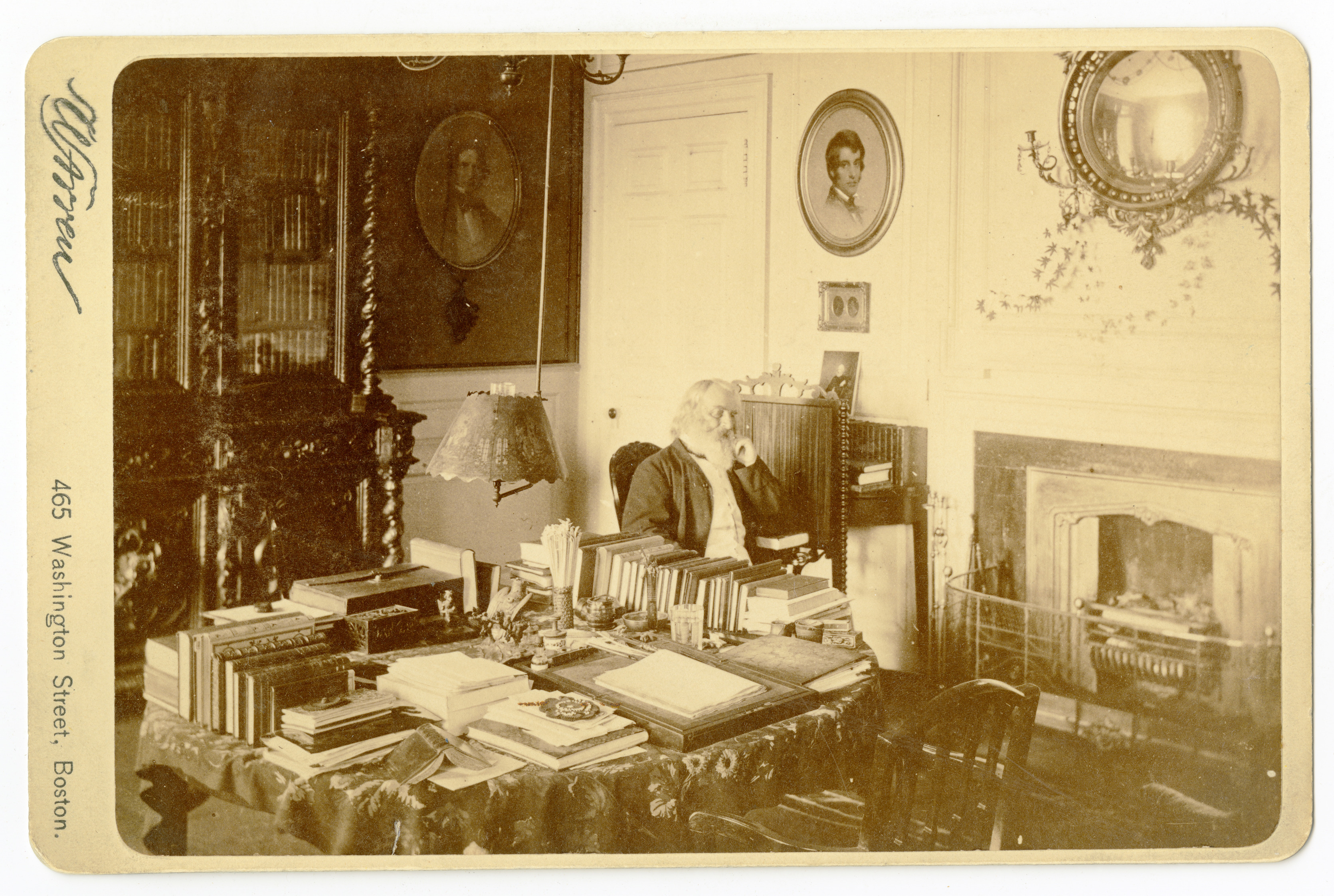 Black and white photograph of bearded old man by fire place in 19th century study.