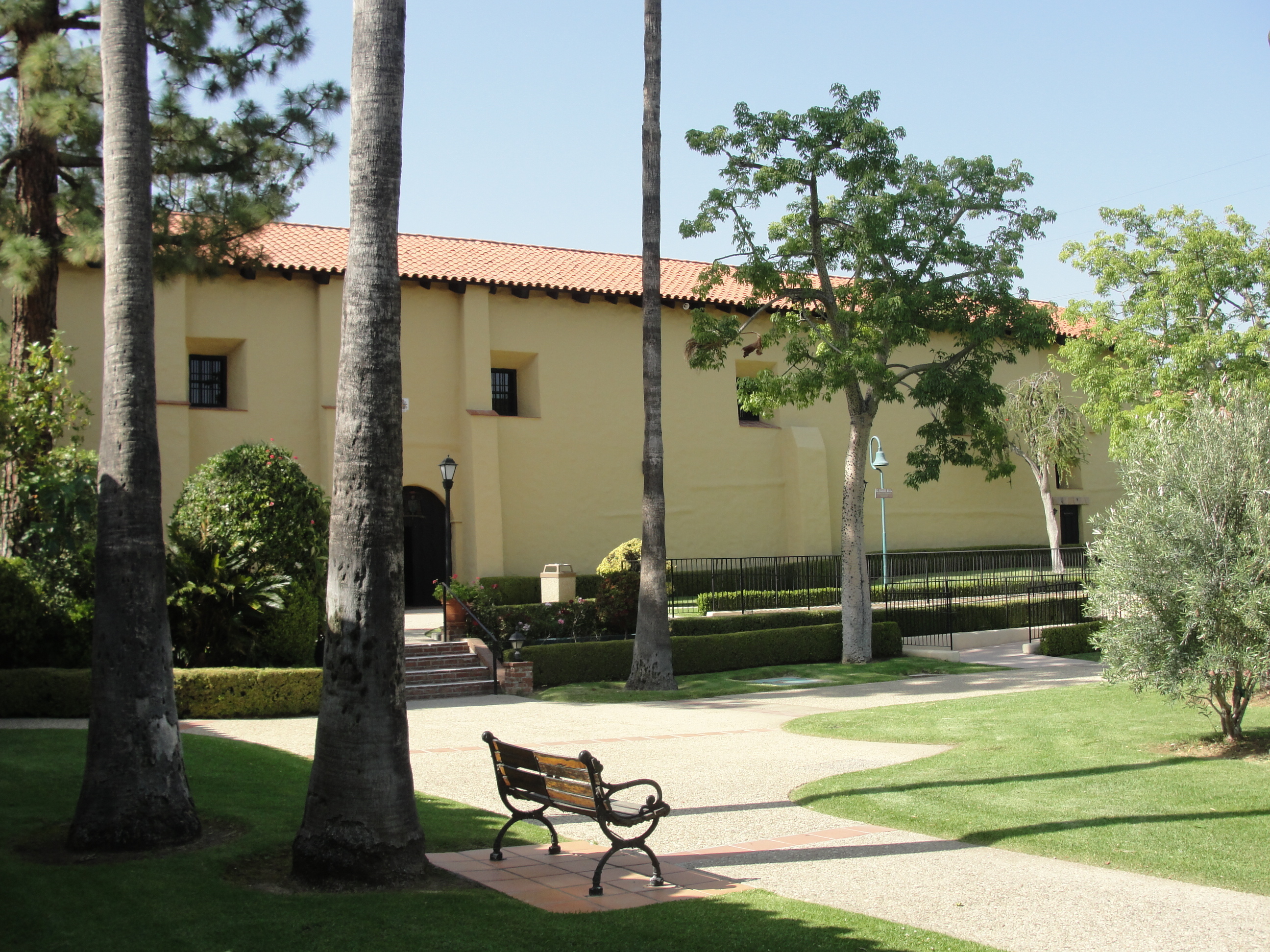 The front of a white mission building with a Spanish-style tiled roof opens to a courtyard garden with mature palm trees, a concrete walkway, and a bench
