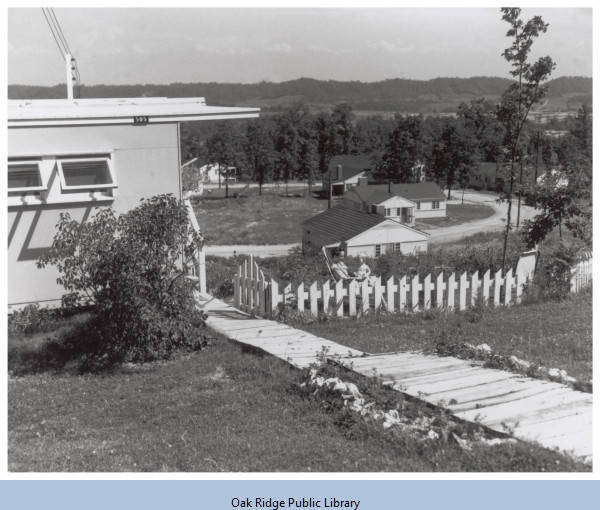 A woman and a child are sitting in a fenced yard of a flattop house. Other houses are in the distance.