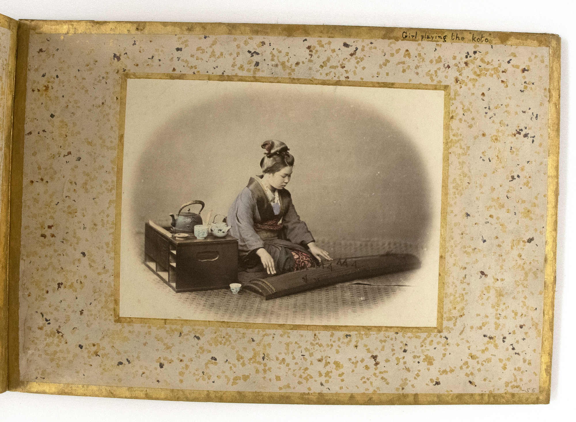 Sepia photograph colored with blue, pink, yellow, purple and red shades representing a Japanese woman sitting on a mat by a box with tea accessories and playing musical instrument placed on the floor. She is photographed against a plain wall in the background. The photograph is placed in the center of an album page decorated with a gold-leaf frame, colorful dots, and an inscription.