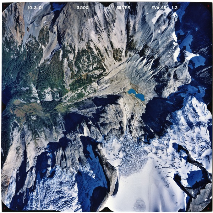 Color aerial photograph of glacier and snowfields on mountain peak with forested valleys below.