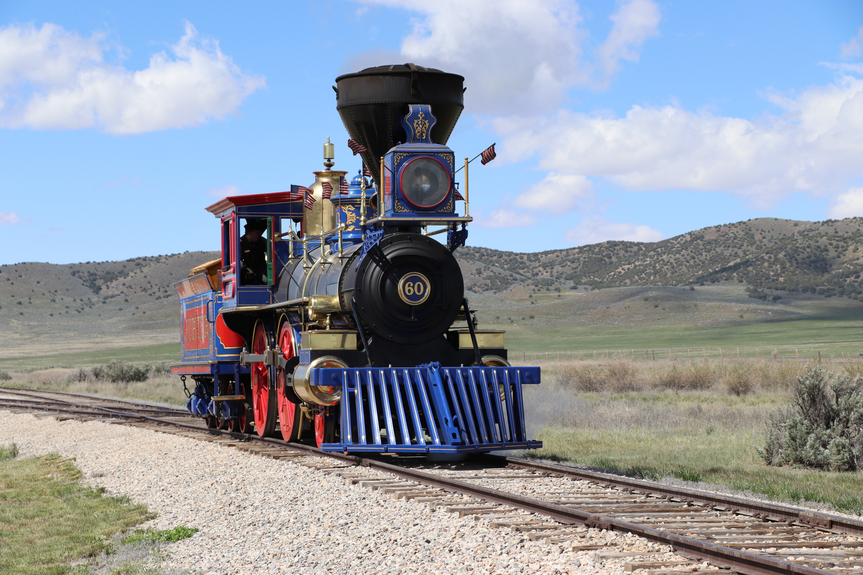 Blue, red, black and gold steam locomotive sits on a railroad track. Small American flags fly from the top of the engine. Mountains, sky, and sagebrush desert are visible in the background.
