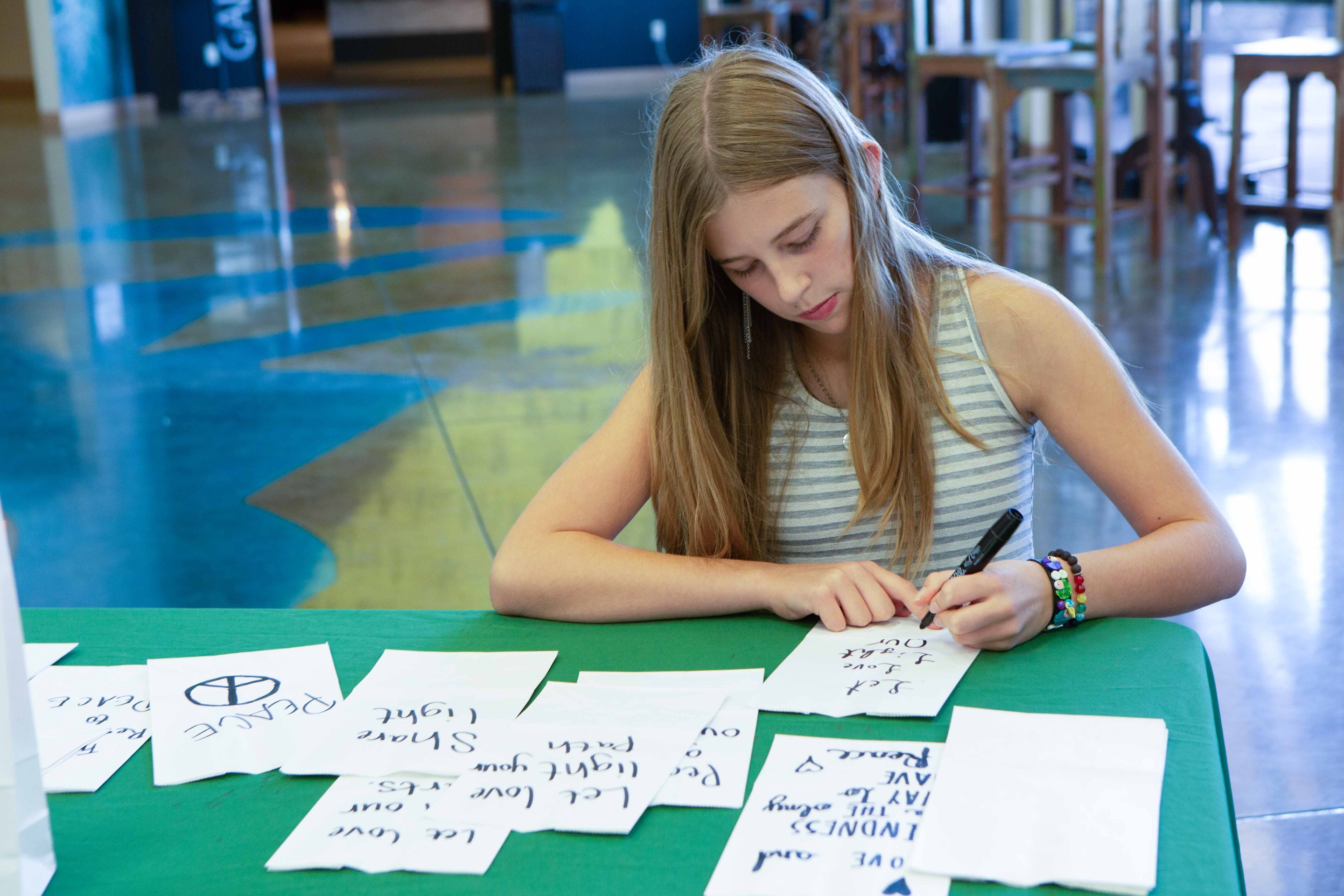 A young person writes a message on a white bag on a table of other white bags with messages of peace. 
