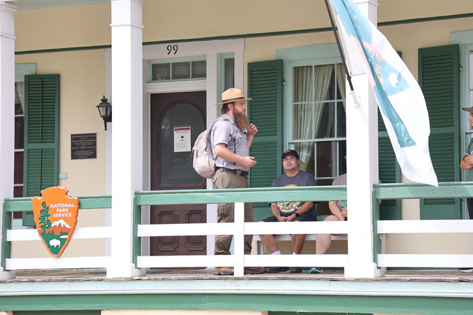 Park Ranger standing on white and green accented porch of a yellow home. NPS arrowhead and park service flag are on display as he speaks to a small group of visitors.