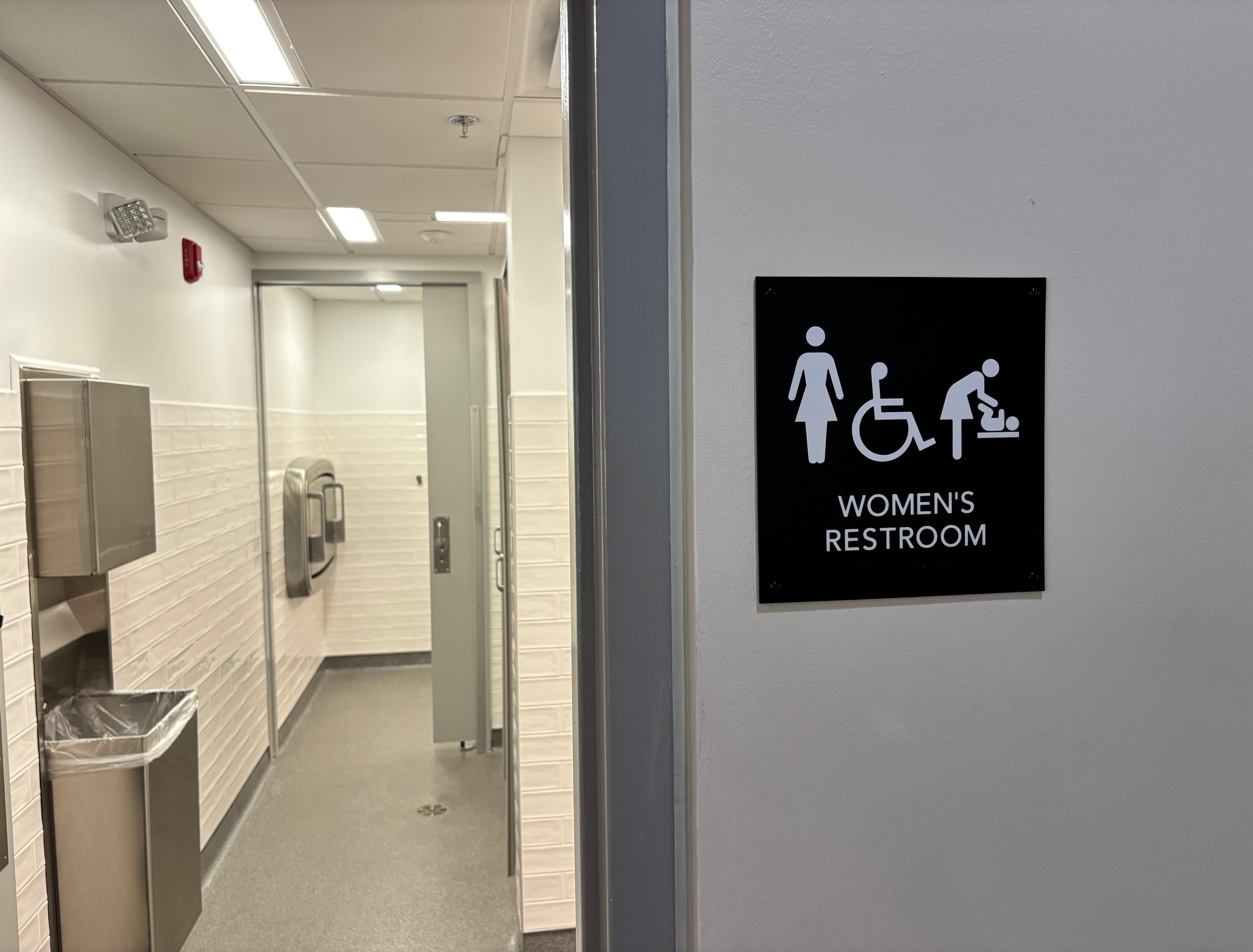 A look into a long bathroom with several stalls to the side and a handicap stall furthest back. New, white tile decorate the walls.