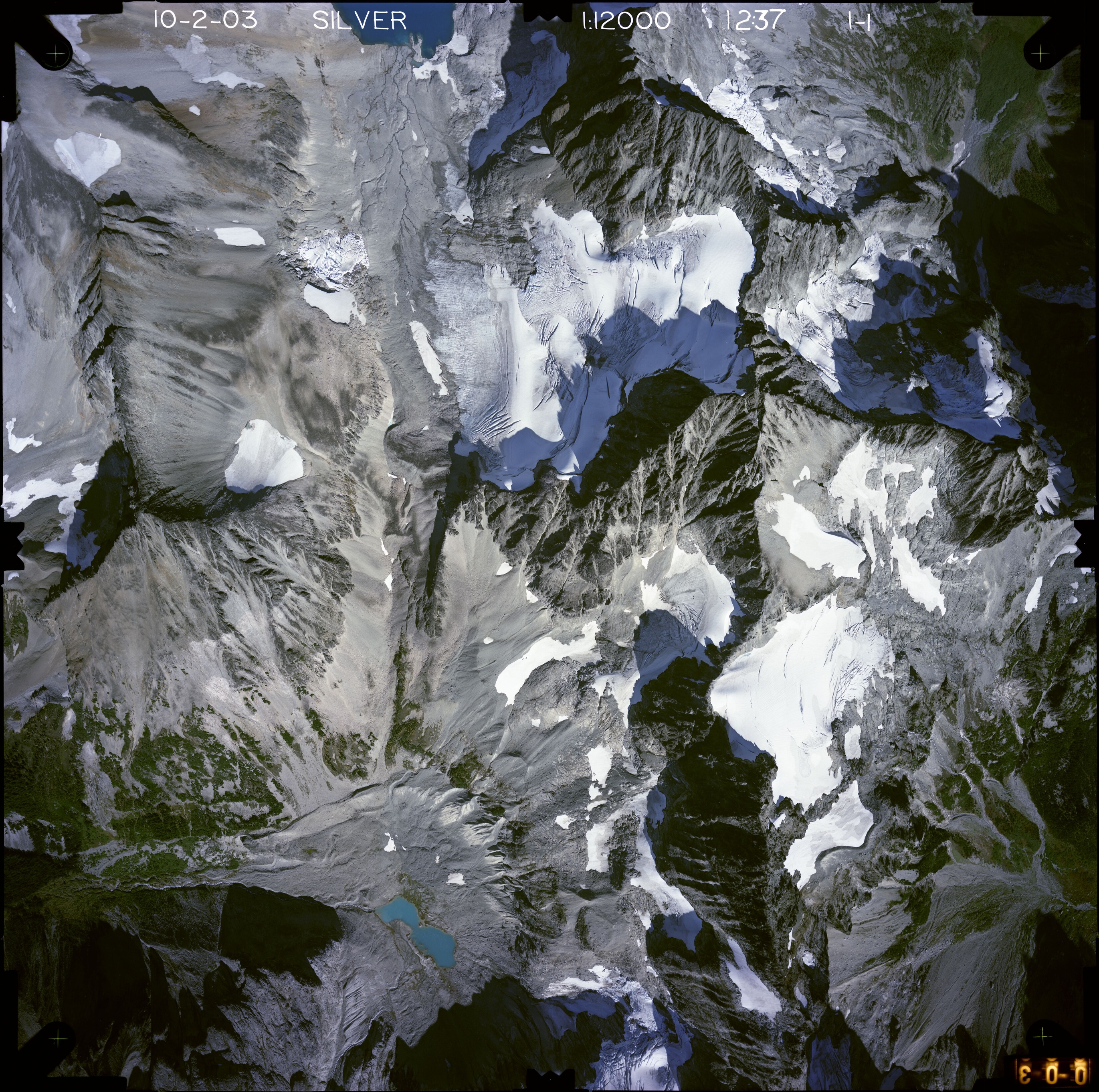 Color aerial photograph of glacier, snowfields, and alpine lake on mountain peak with forested valleys below.