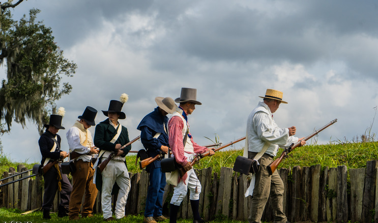 Outside daytime. 6 men in period clothing stand together in a line by a wooden fence. They are holding guns.