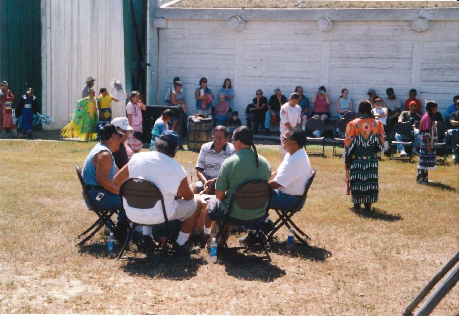 A group of men sitting around a large drum with girls in American Indian Dress Dancing behind them