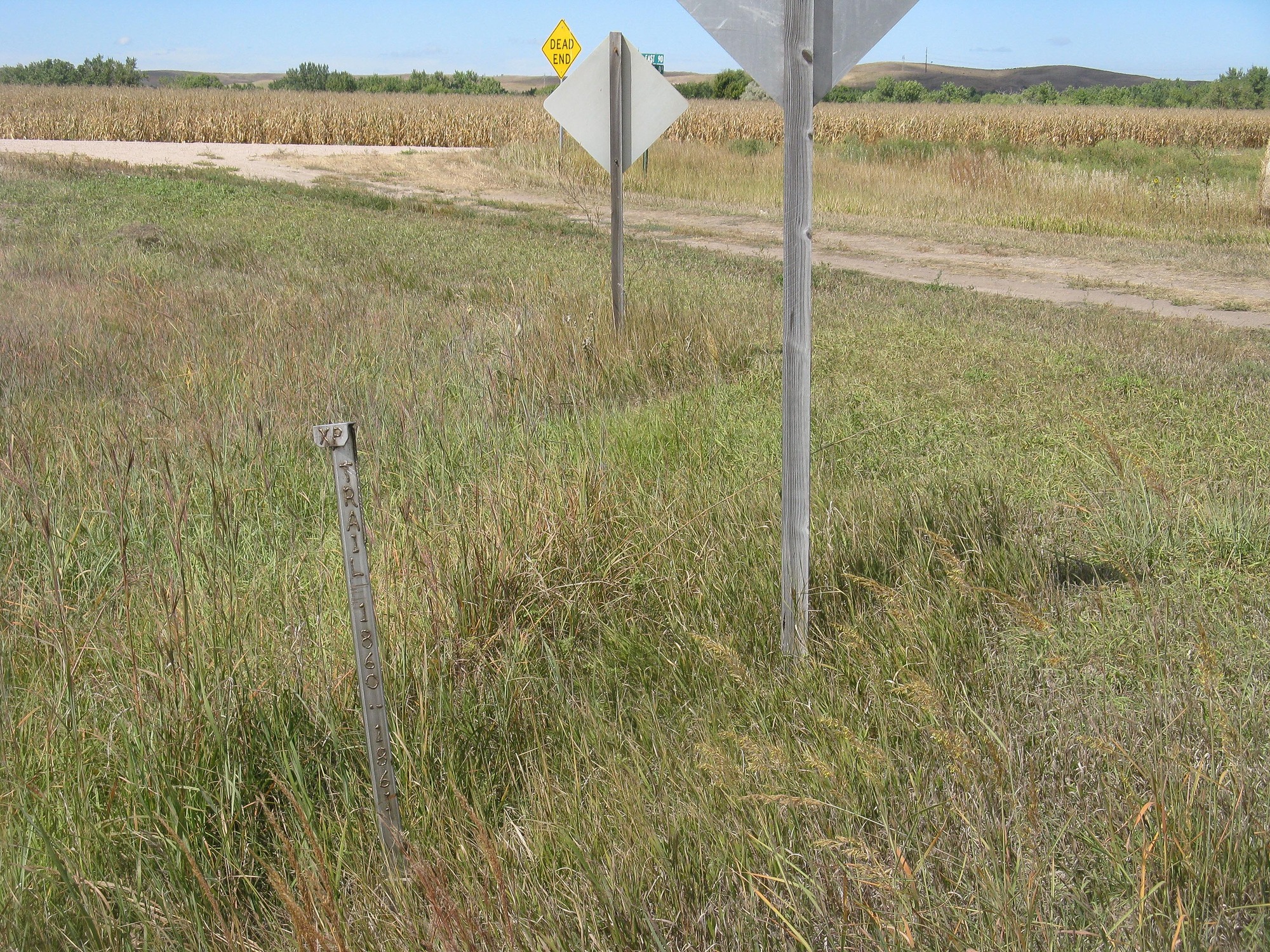 A metal pole marking the location of Gills Pony Express station rises out of a field of grass next to a dirt road and crop fields