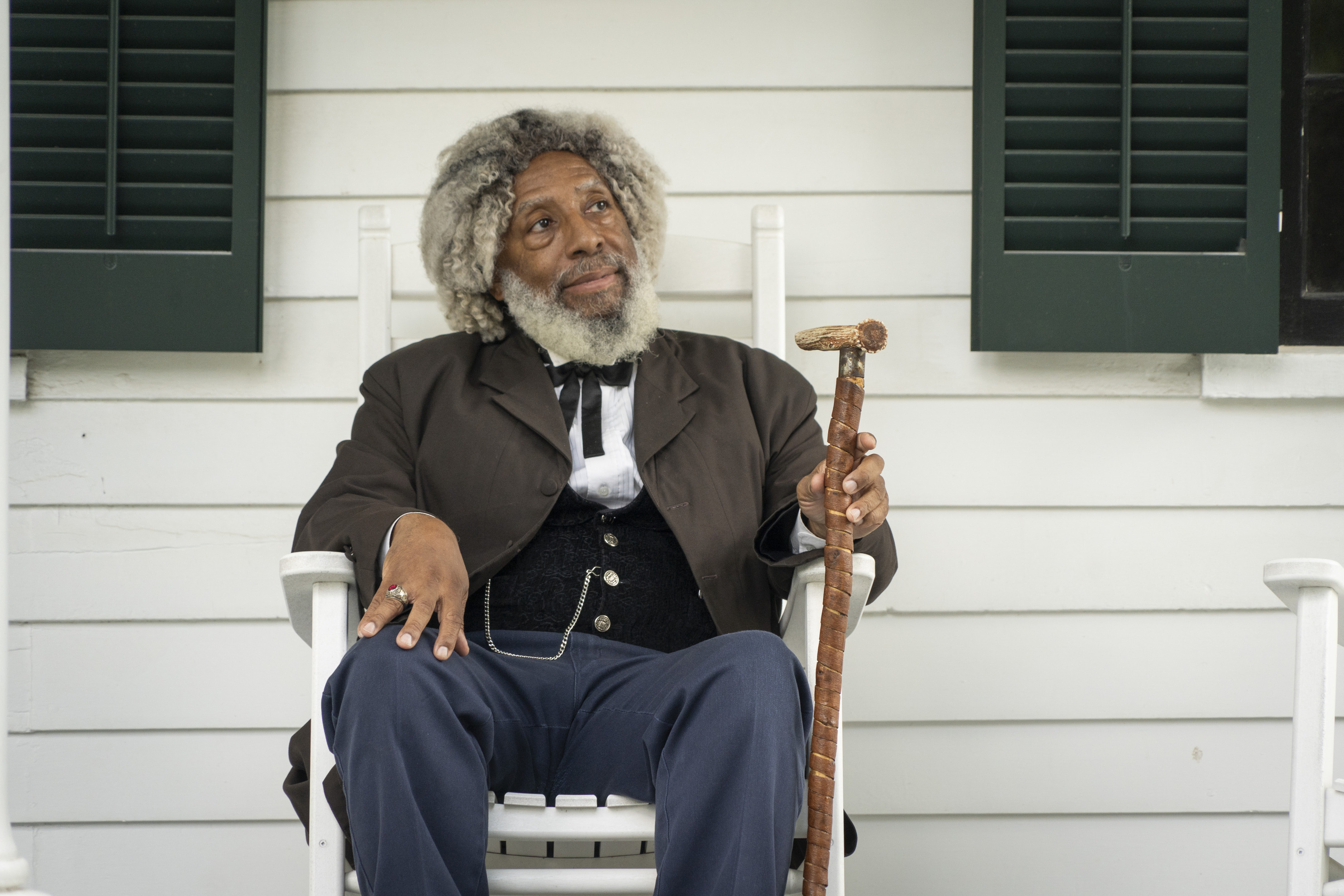 A man dressed in 19th century clothes with a cane sits in a rocking chair on a porch.