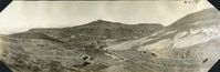 This is an historic black and white, panoramic photograph from the Scotty's Castle Historic Photograph Collection, Death Valley National Park with expansive view of desert hillsides and canyon with building complex in wash. Primary structures nestled in middle. Temporary structures making up Indian Camp along newly graded dirt road to left of center. Stacks of railroad tiles line dirt road to right. Desert hillsides should geological process. Other mountains faint in the distance.
