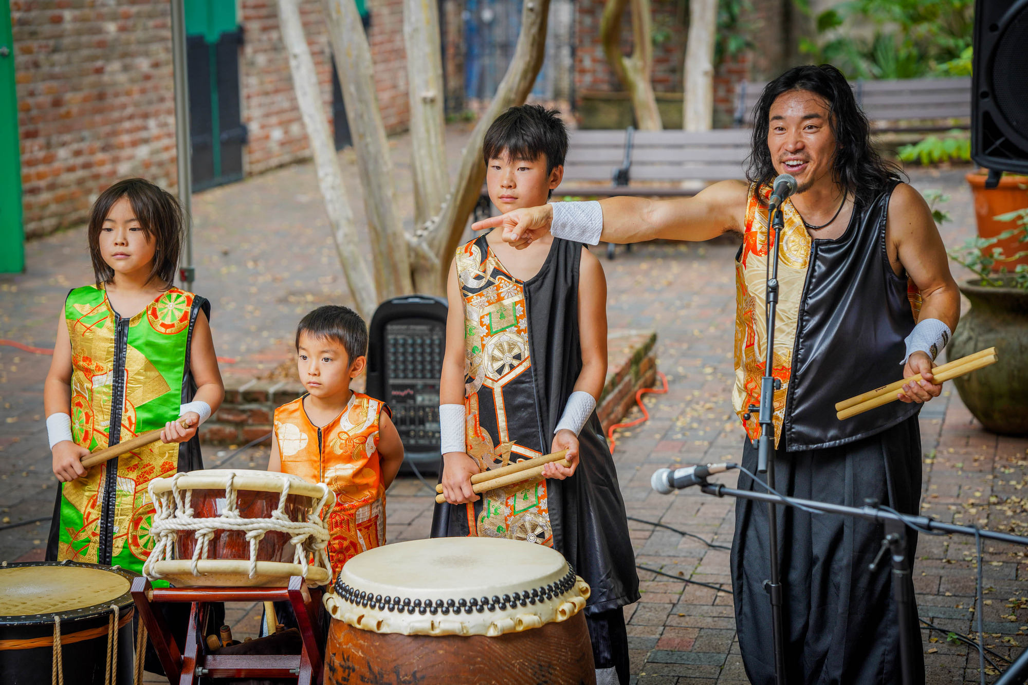 Four percussionists standing behind drums while one is talking into microphone in a courtyard setting