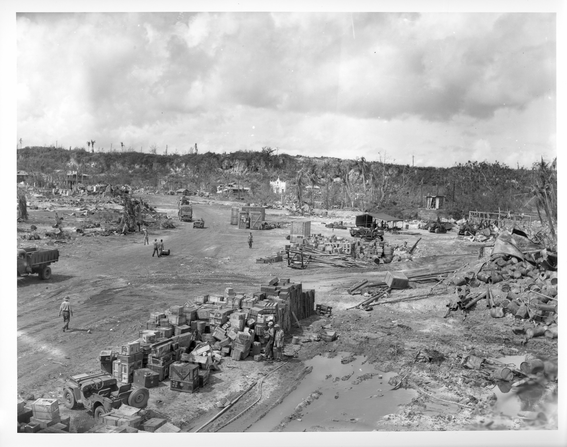 Black and white photo of a ruined town full of piles of boxes being unloaded.