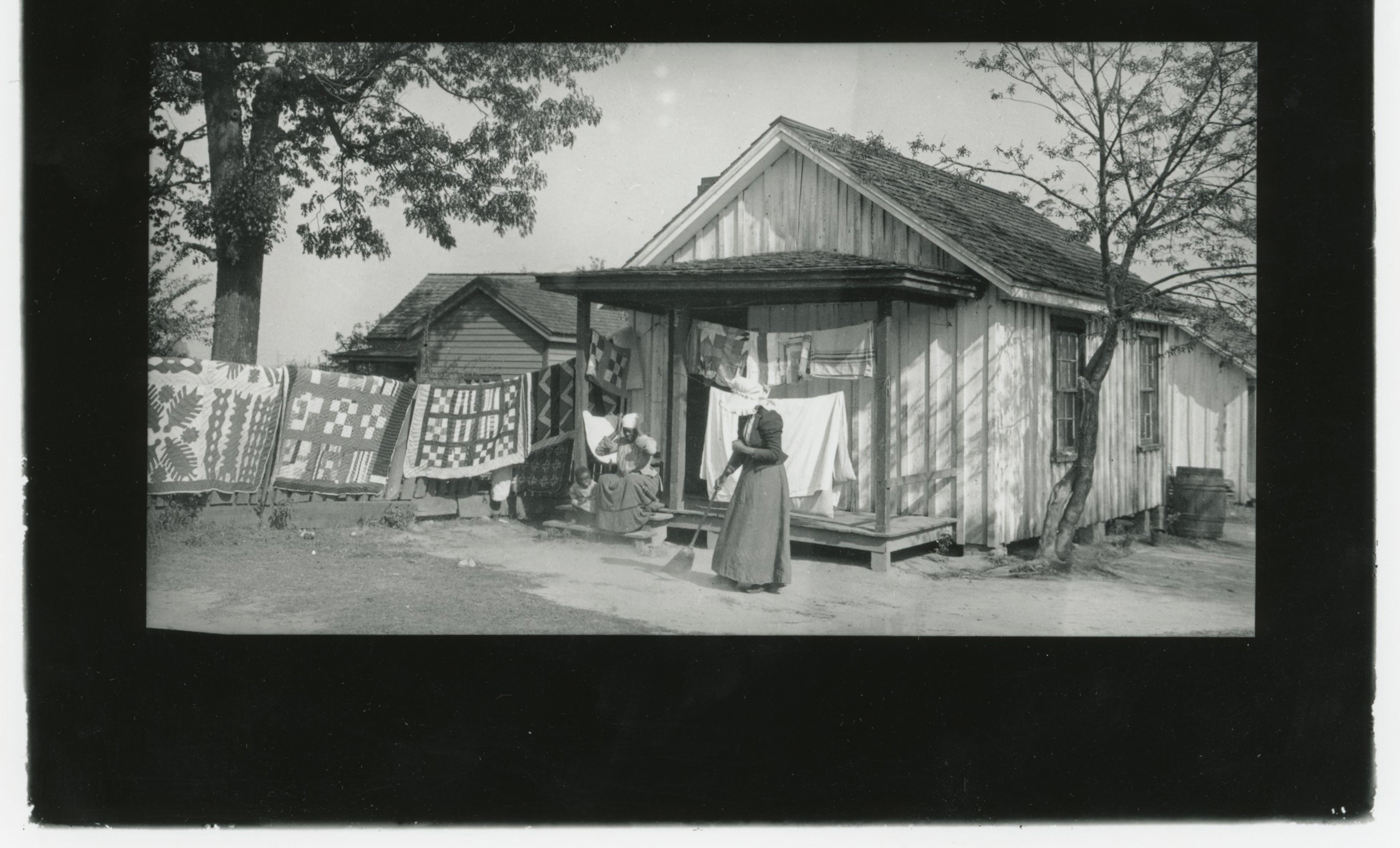 African American woman in white bonnet sweeps ground in front of one-room house. African American woman and child sit on front porch. Boldly patterned quilts hang on clothesline on the left of the image.