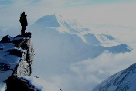 person on a rocky outcropping, looking at a massive snow-covered mountain