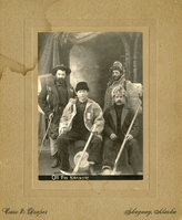 Black & white albumen print of four greenhorn Stampeders posed in brand-new furs and coats with shiny unused mining pan and picks.  One has a hiking stick and has a box strapped to his back.  This is a posed studio portrait with a painted architectural backdrop that includes a pedestal and curtain to the left side of the image.  The print is mounted in a tan cabinet card with "Case and Draper" printed at the bottom.  "Off for Klondyke" is written at the bottom of the albumen print, probably made by scratching into the negative prior to printing.  